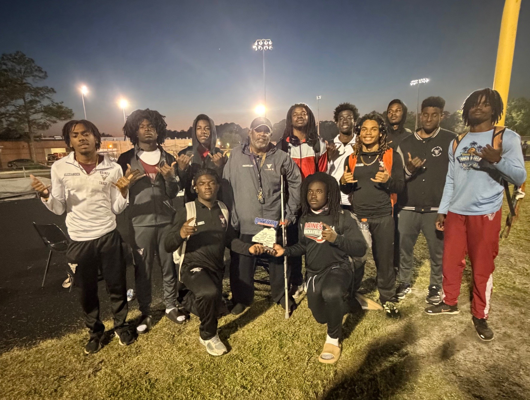 A group of people, some in sports attire, stands outside on grass under night lights. They are smiling and posing for a photo.