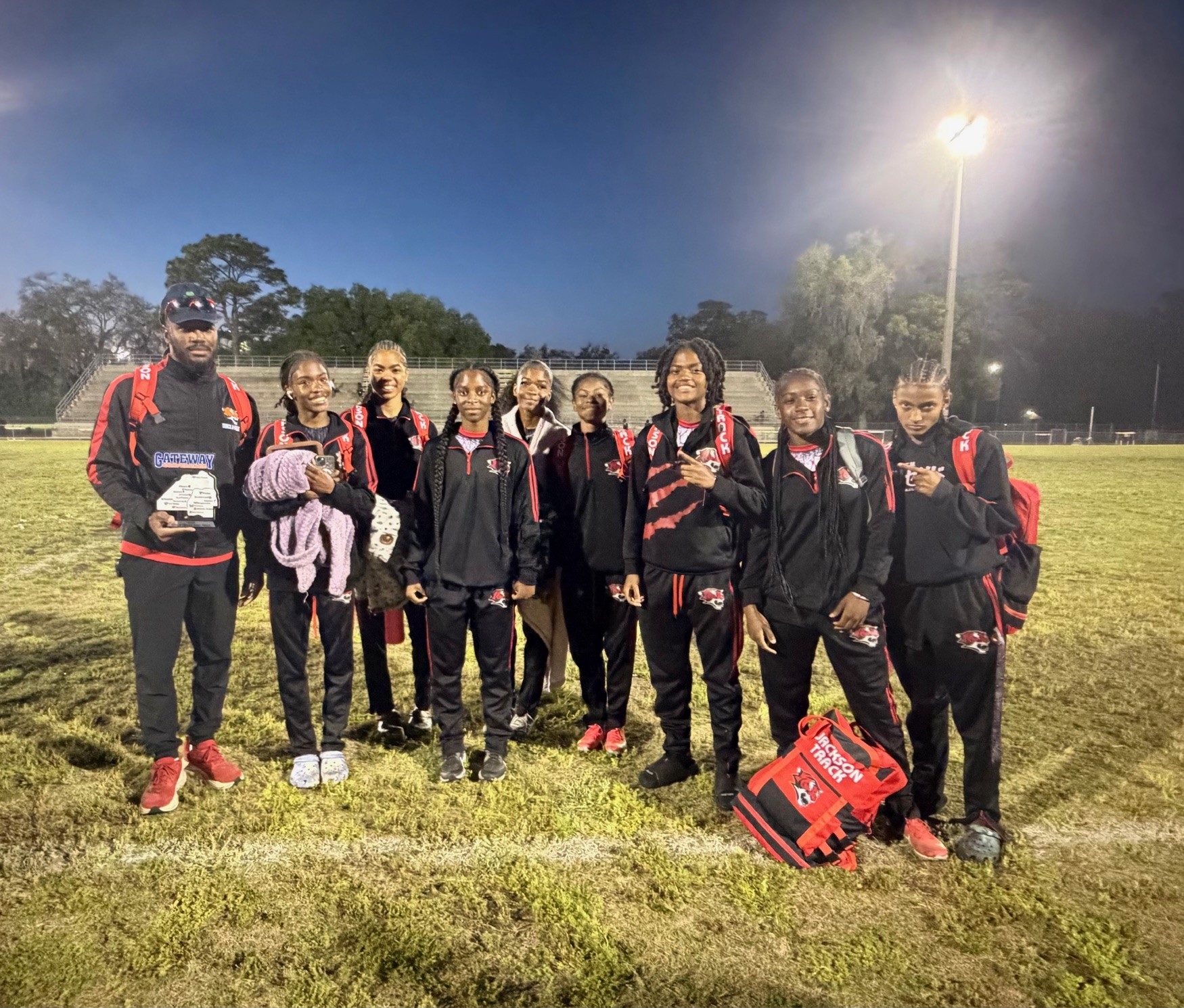 A team of athletes wearing black and red uniforms stand together on a field at night.