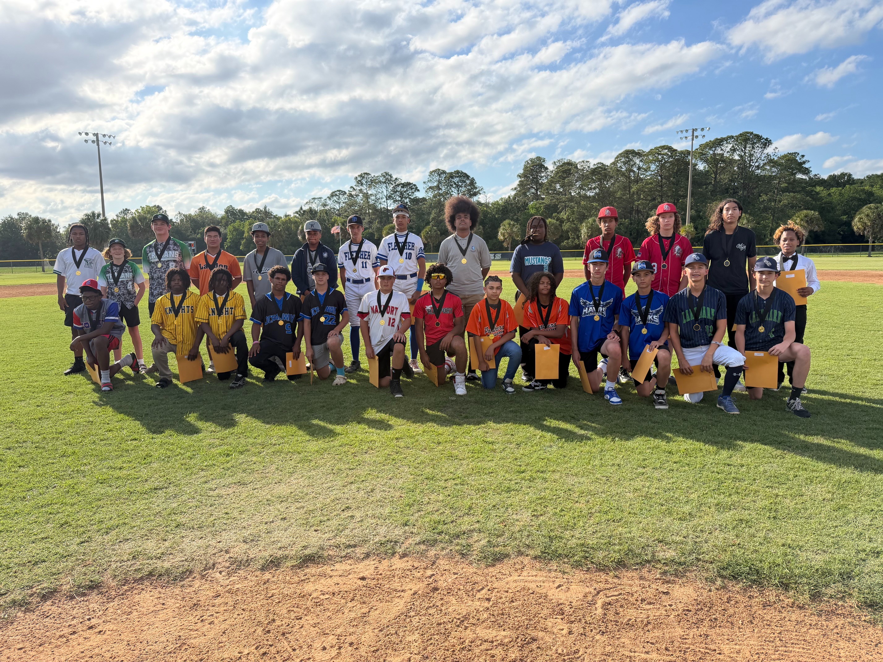 Group photo on a baseball field. Many people in sports uniforms, some with medals, on a grassy field.