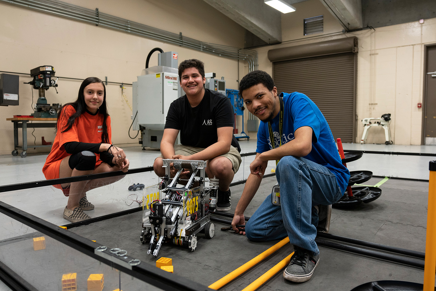 Three students in casual clothes, two kneeling and one sitting, around a robot on the floor.