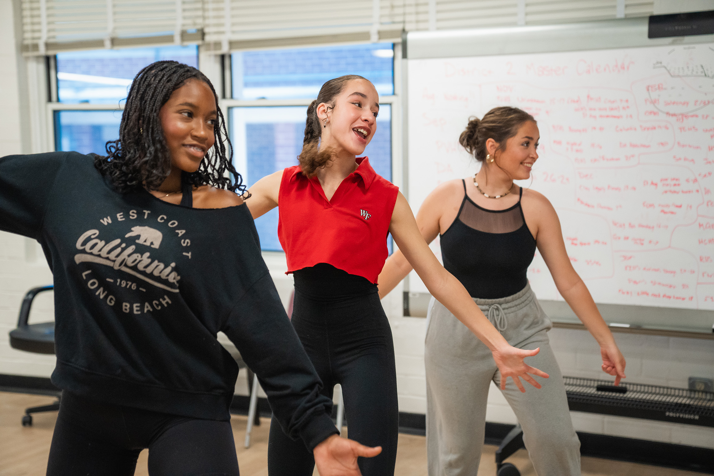 Three women in a dance studio, two smiling, one laughing, wearing various colored tops and leggings, and a whiteboard in the background.