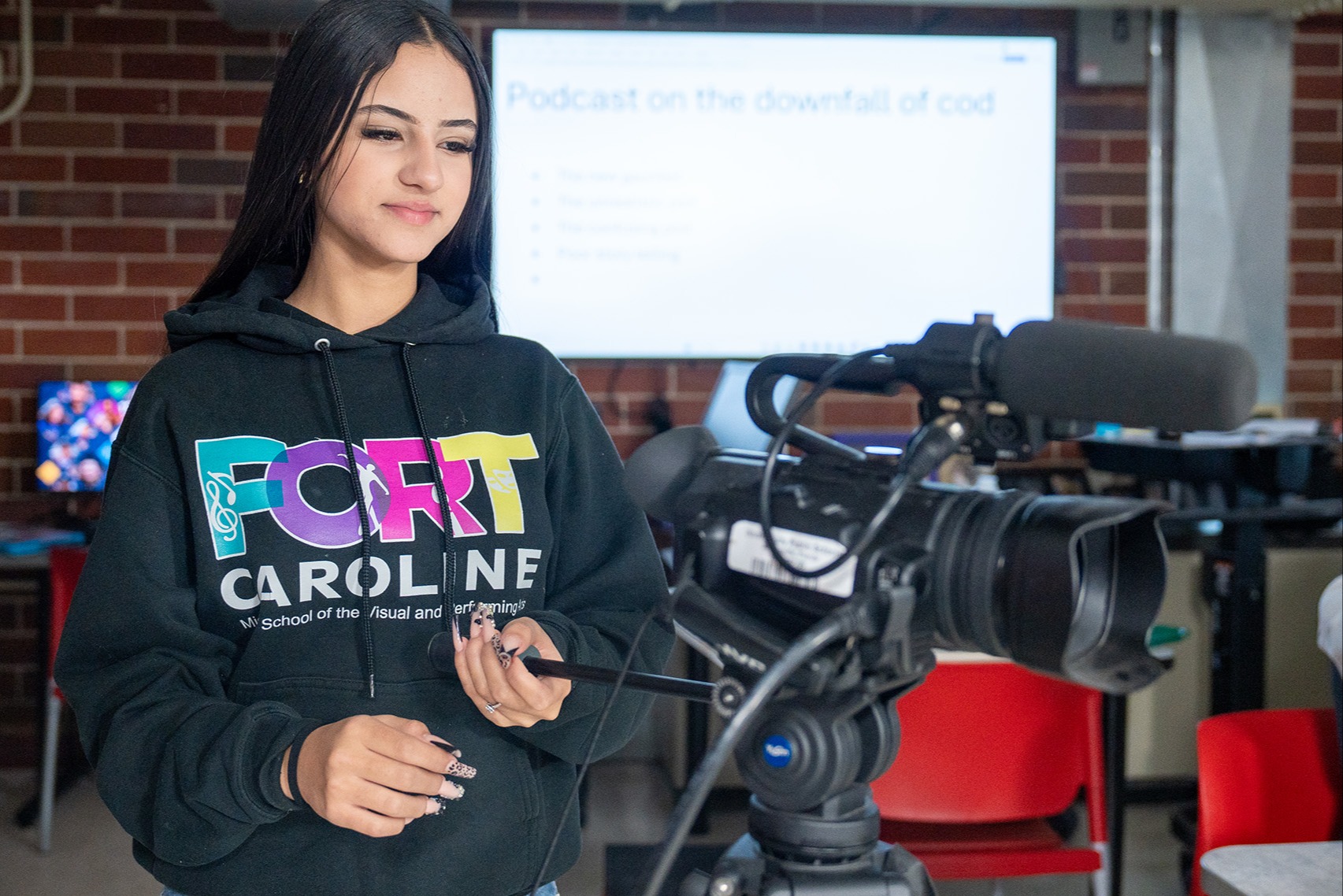 A woman wearing a colorful hoodie stands in a classroom, holding a microphone and camera. Behind her is a projector screen and brick wall.