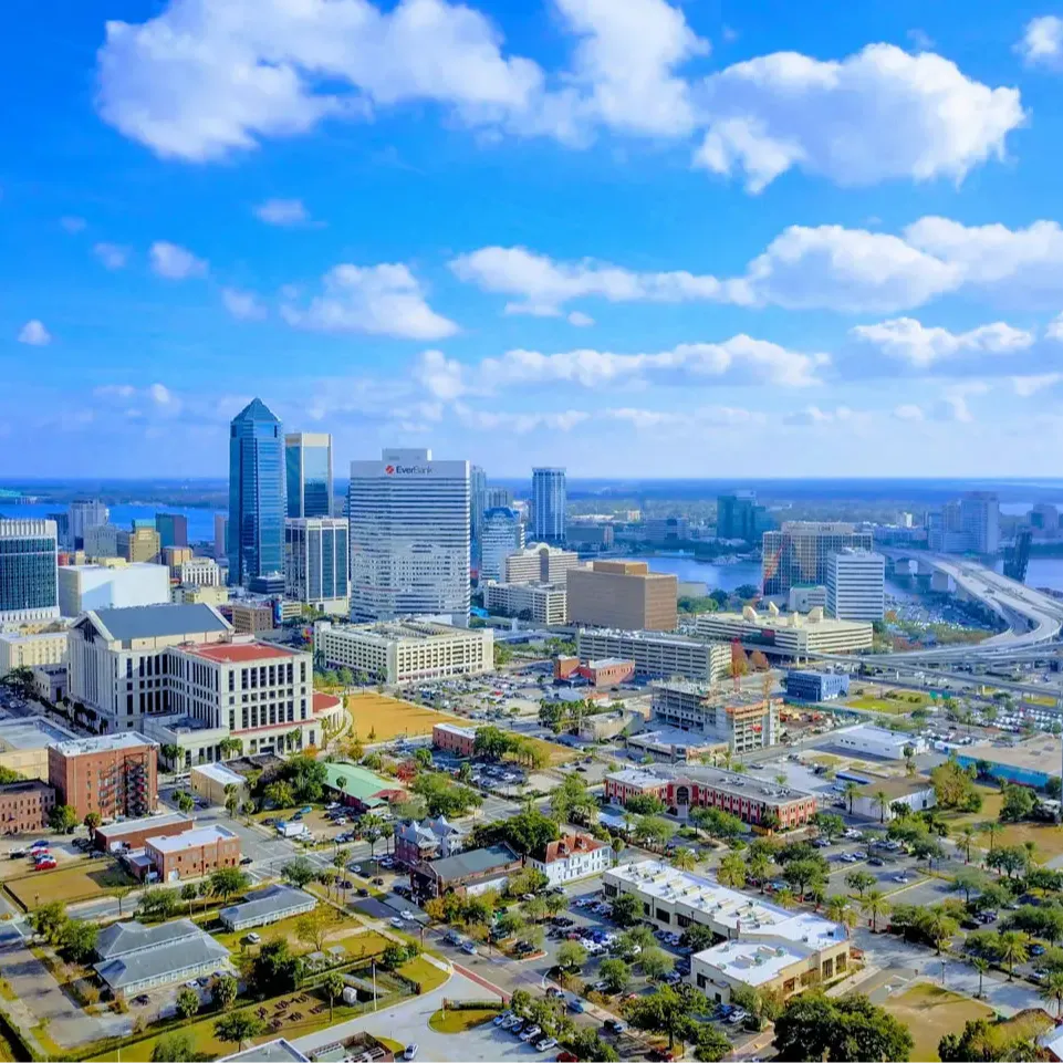 Aerial view of a city with tall buildings, a river, and a bridge. The sky is blue with white clouds.