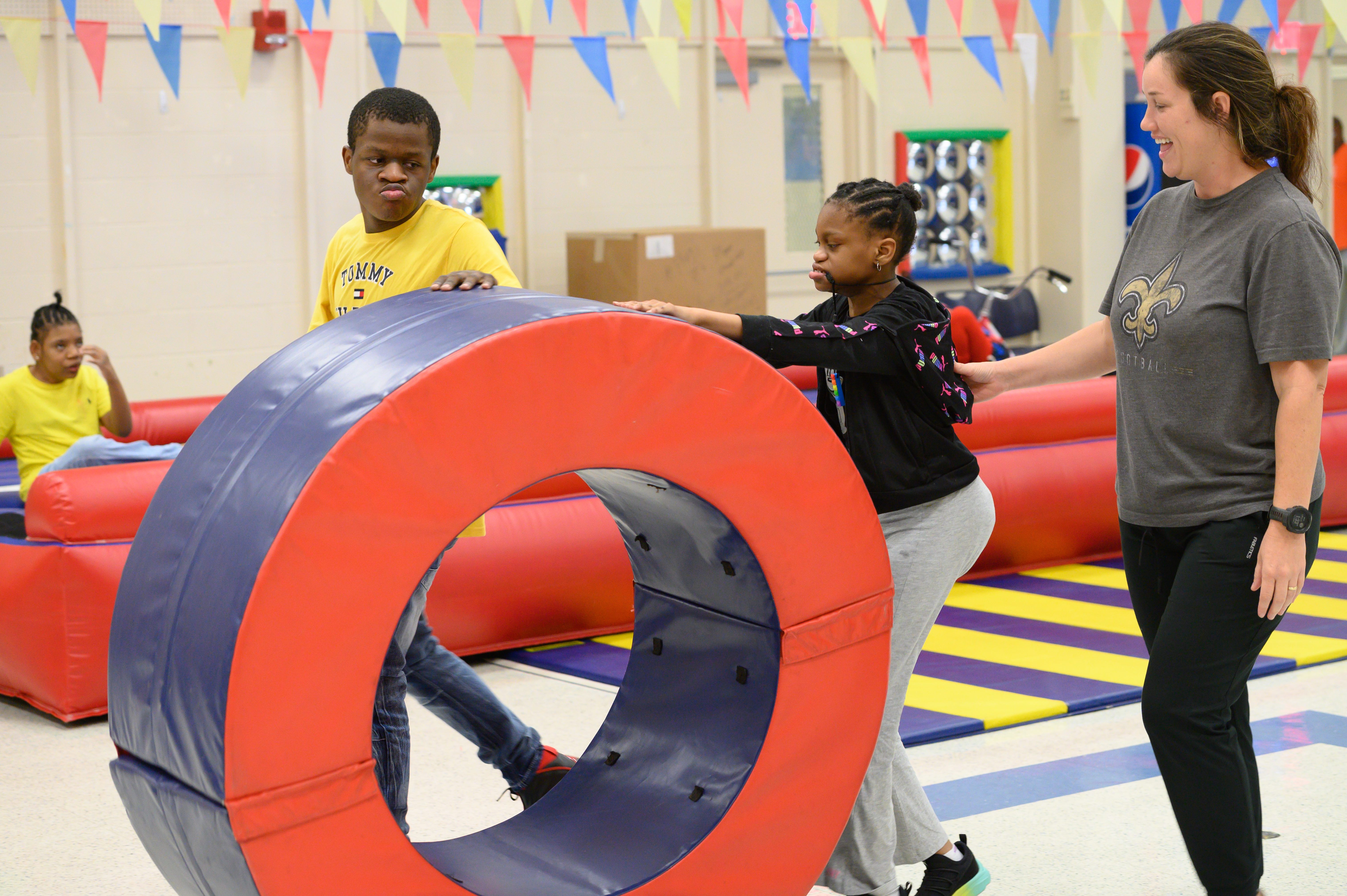A woman guides a child through a large red and blue ring in a playroom with other children.