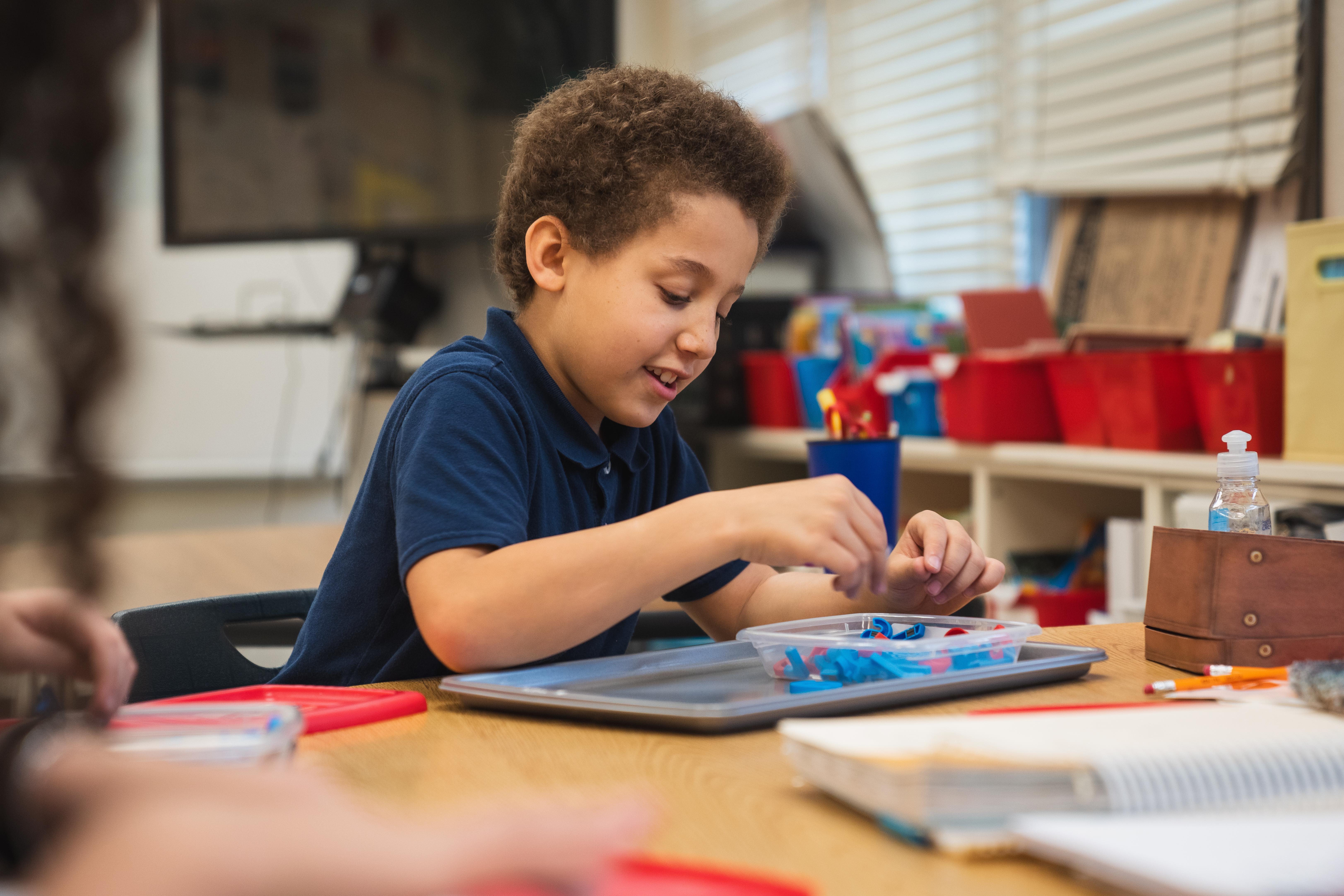 Child seated at a table, focused on a craft project with a teacher assisting in the background.