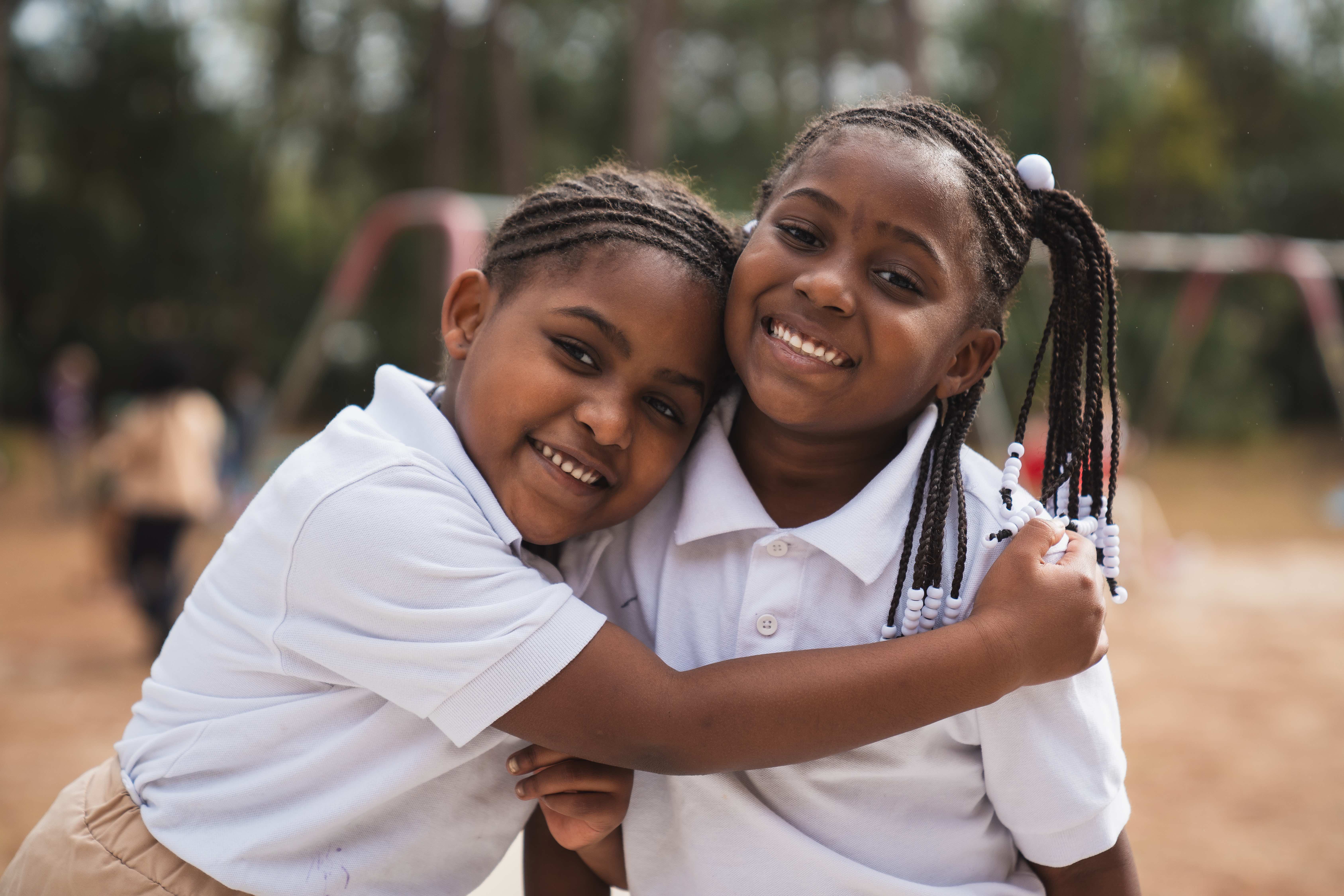 Two children in white shirts embrace and smile in an outdoor setting with playground equipment in the background.