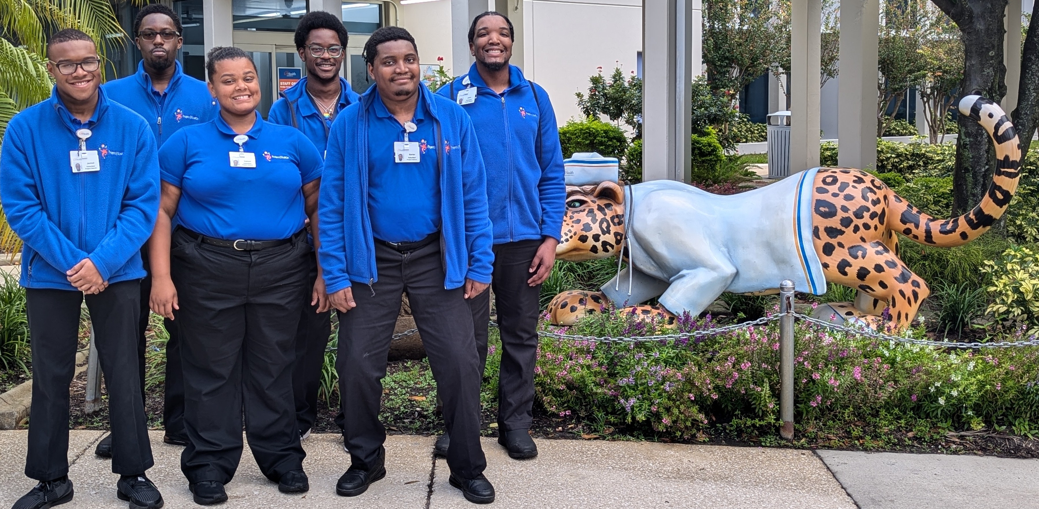 A group of people in matching blue jackets stand next to a large leopard statue in front of a building.