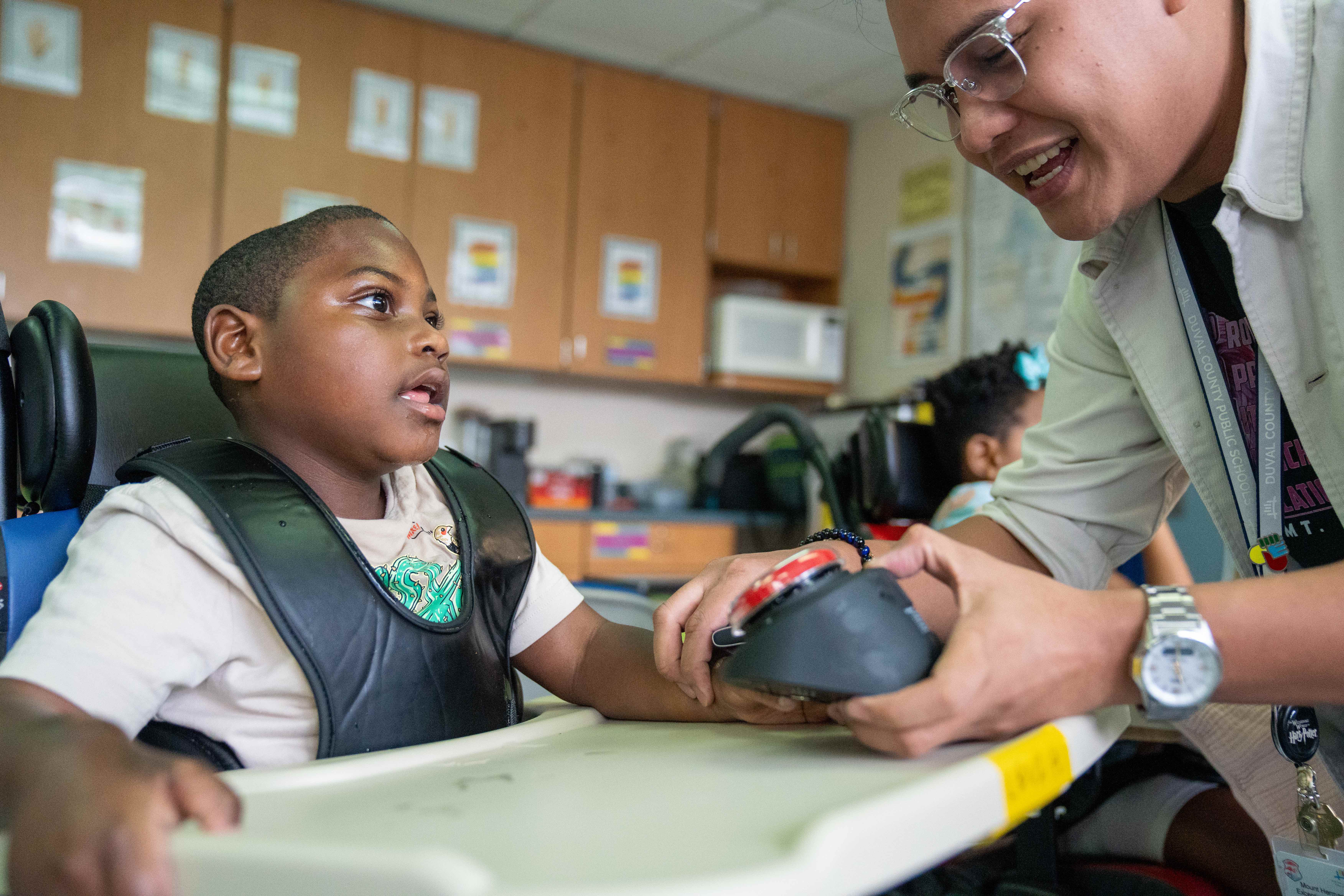 A child in a wheelchair reaches for a device held by a smiling adult, in a classroom with wooden cabinets and a microwave.