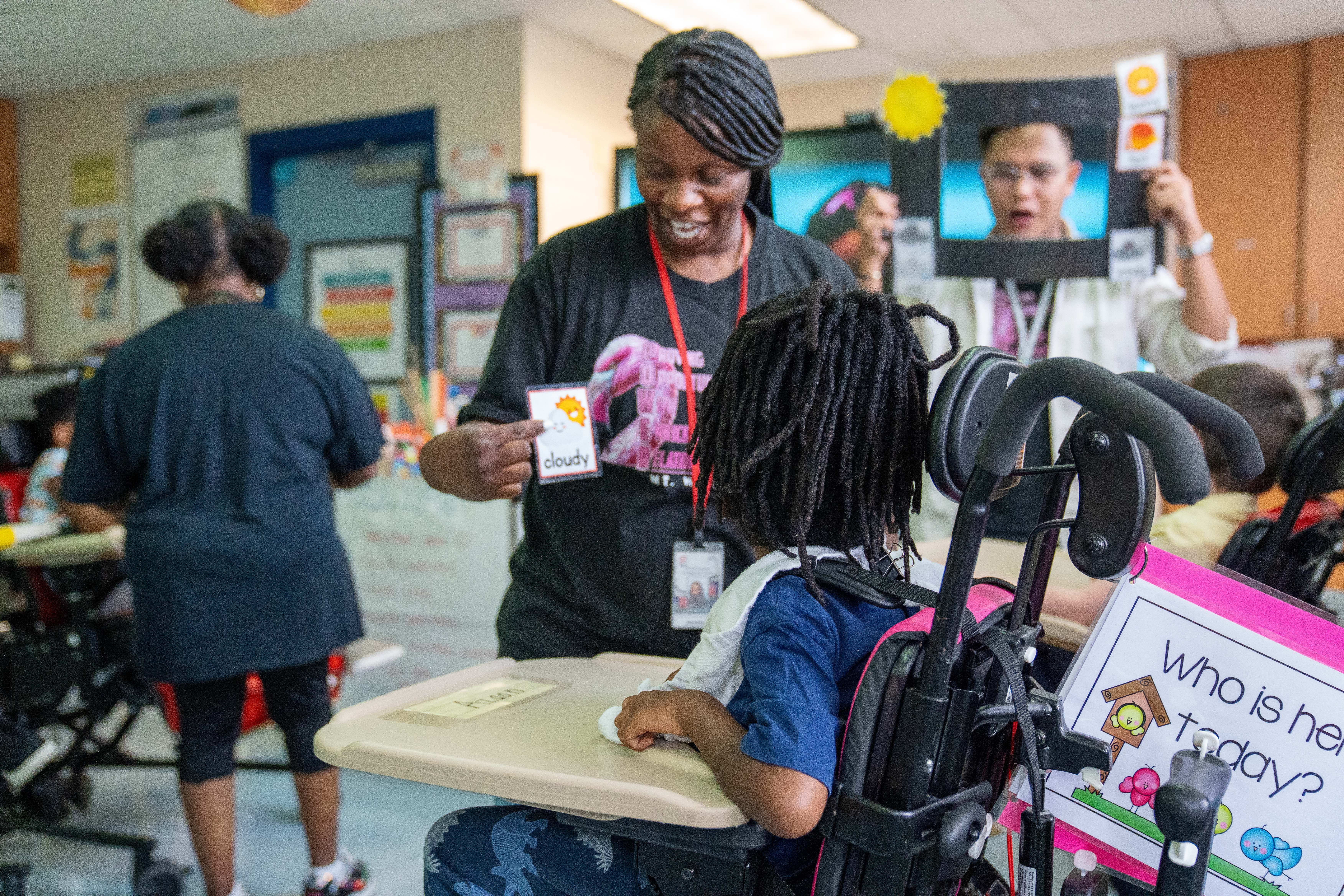 A teacher in a classroom helps a student in a wheelchair. Other students are in the background.