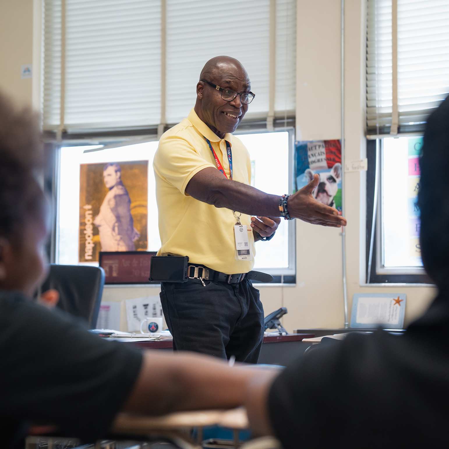 A man in a yellow shirt gestures while teaching a class; two students, one with an afro, listen.