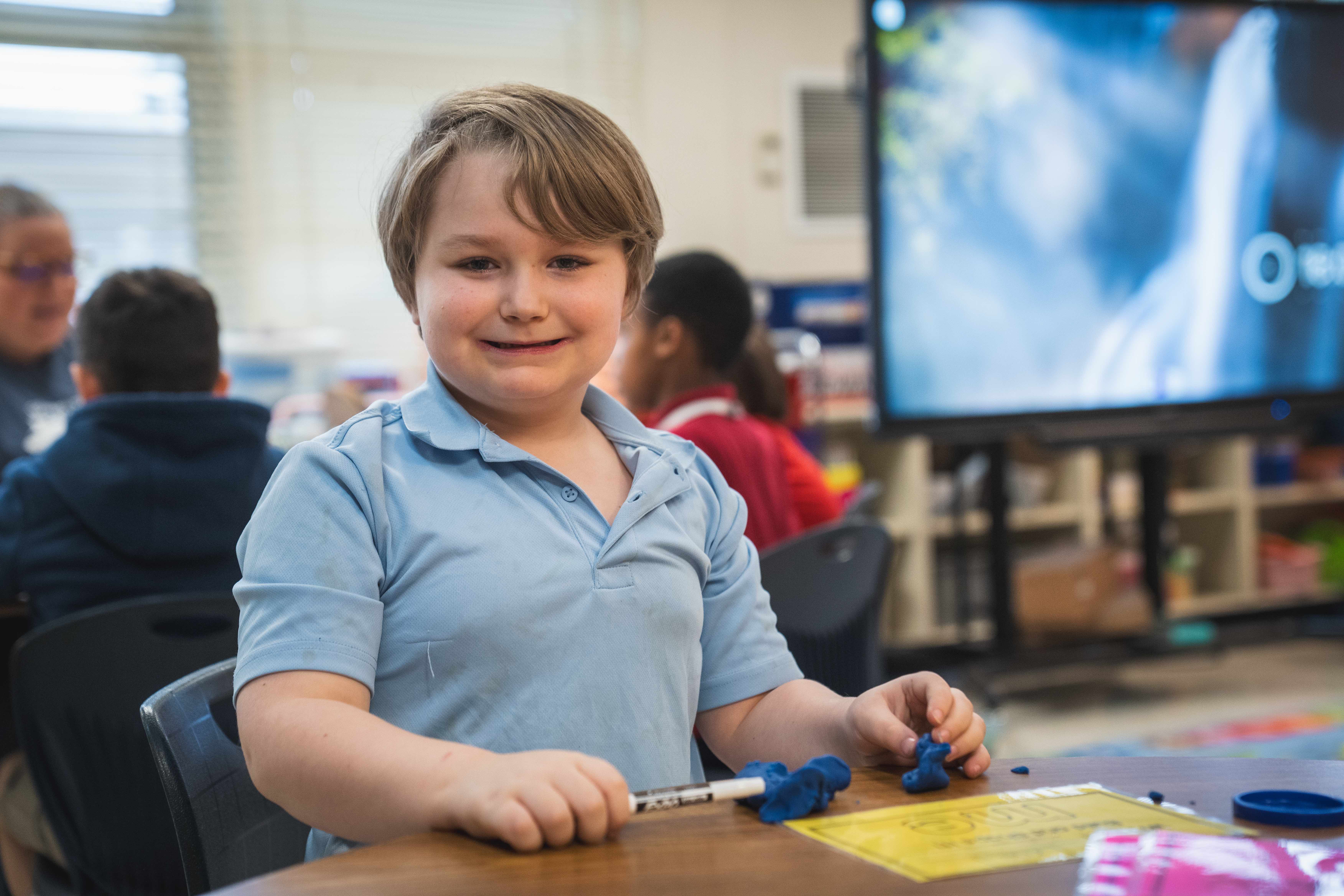 A child wearing a blue shirt is smiling and holding a blue object in a classroom.