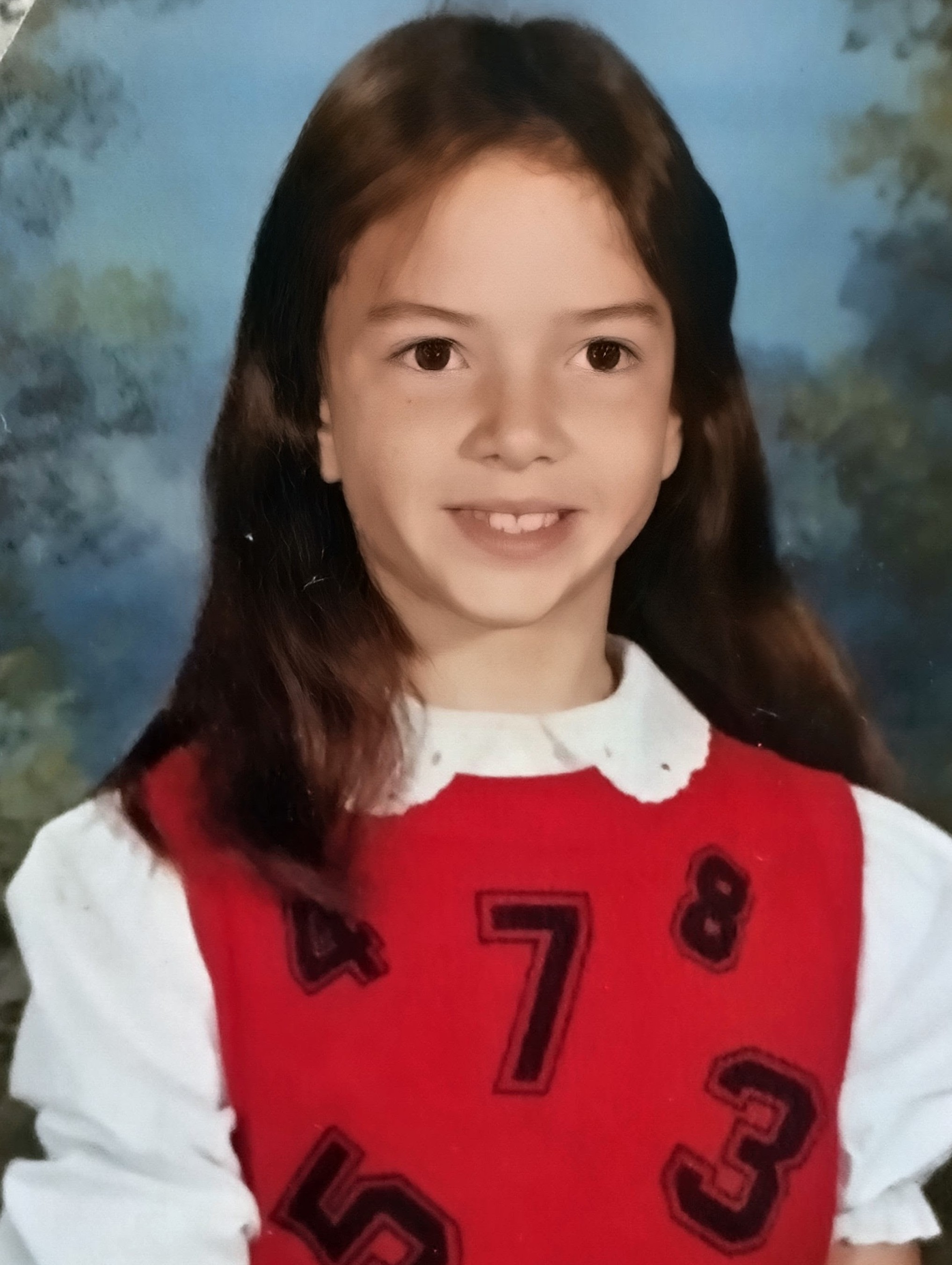 A young girl with long brown hair, smiling, wearing a red and white jersey with numbers 573.
