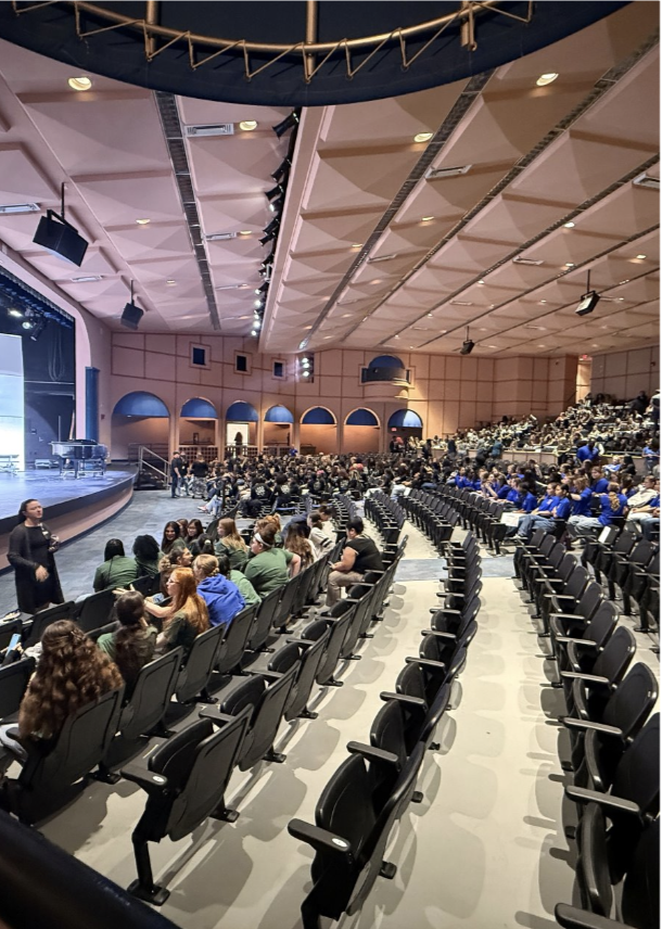 A large auditorium with rows of black chairs, a stage, and many people seated. A person stands at the stage.