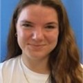 A woman with long hair smiles for the camera, wearing a white shirt. The background is plain blue.