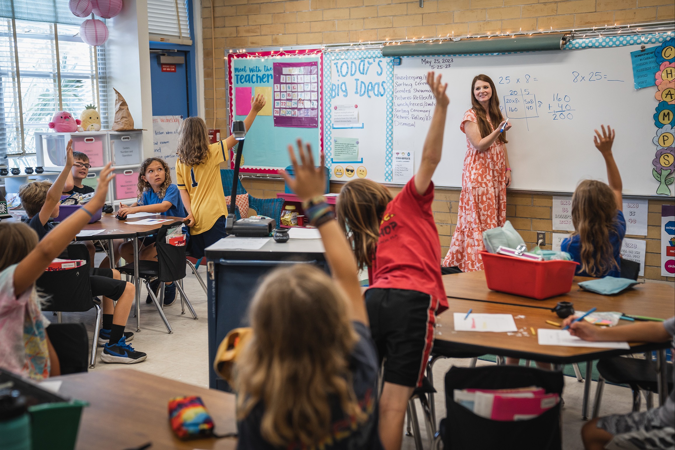 A classroom with children raising hands, a teacher at the front with a whiteboard, and desks arranged in rows.