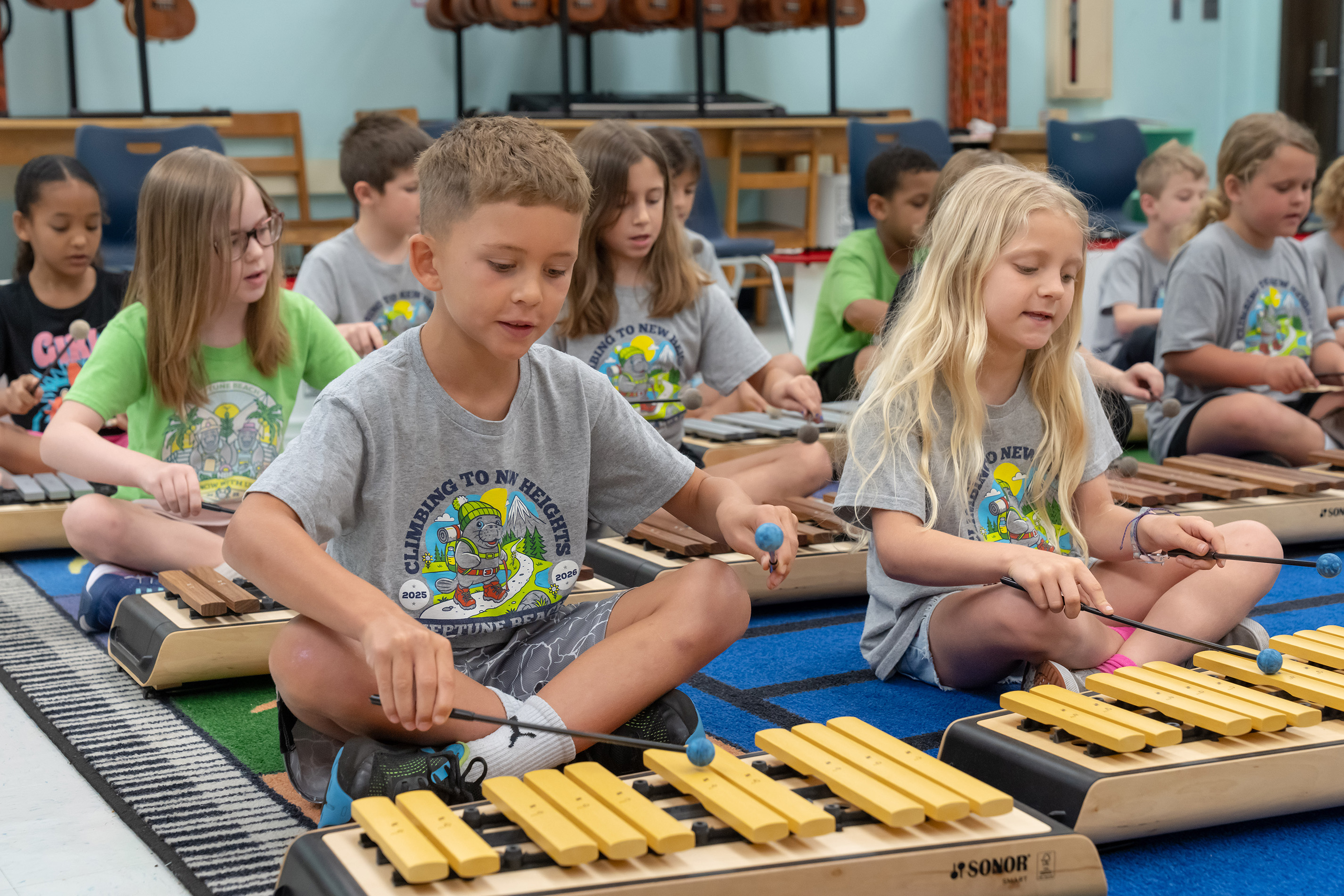 Classroom with children seated on floor, playing xylophones. Background includes wooden chairs and a blue rug.