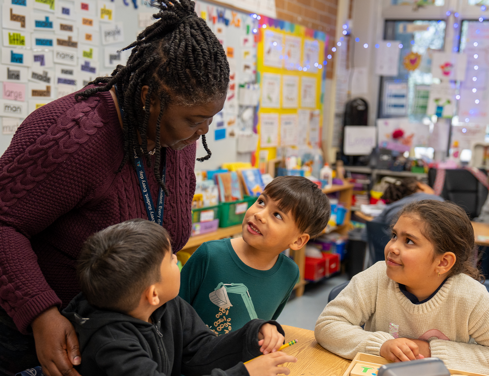 Woman with braided hair helps three young students with school work at a table in a classroom.