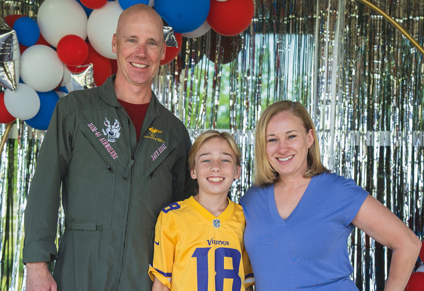Adult man in flight suit, boy in yellow jersey with number 18, woman in blue shirt, posing in front of balloons and silver backdrop.