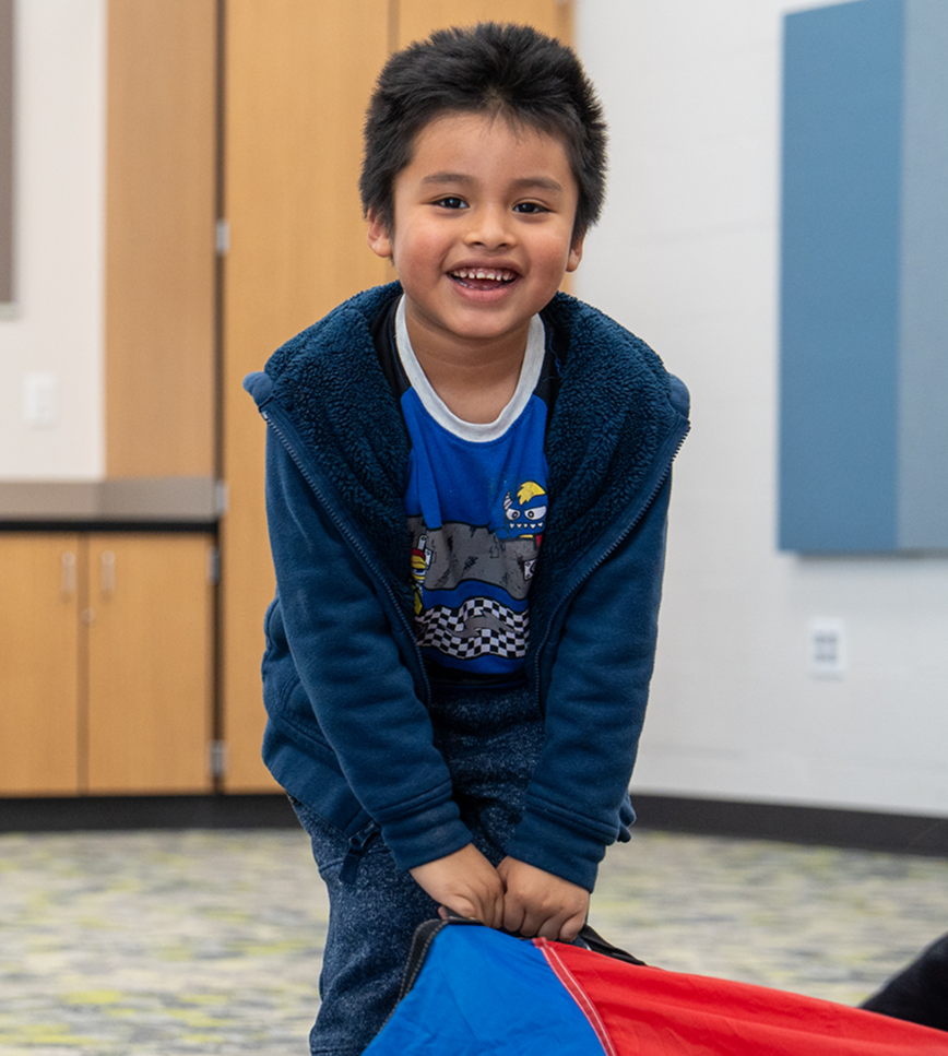 A smiling boy in a blue hoodie holds a colorful kite. Behind him is a room with a blue wall, wooden cabinets, and an open space.