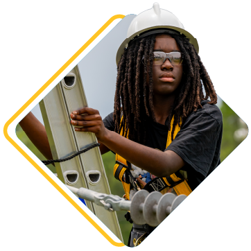 A person in a white hard hat and glasses holds onto a ladder with a utility pole in the background.