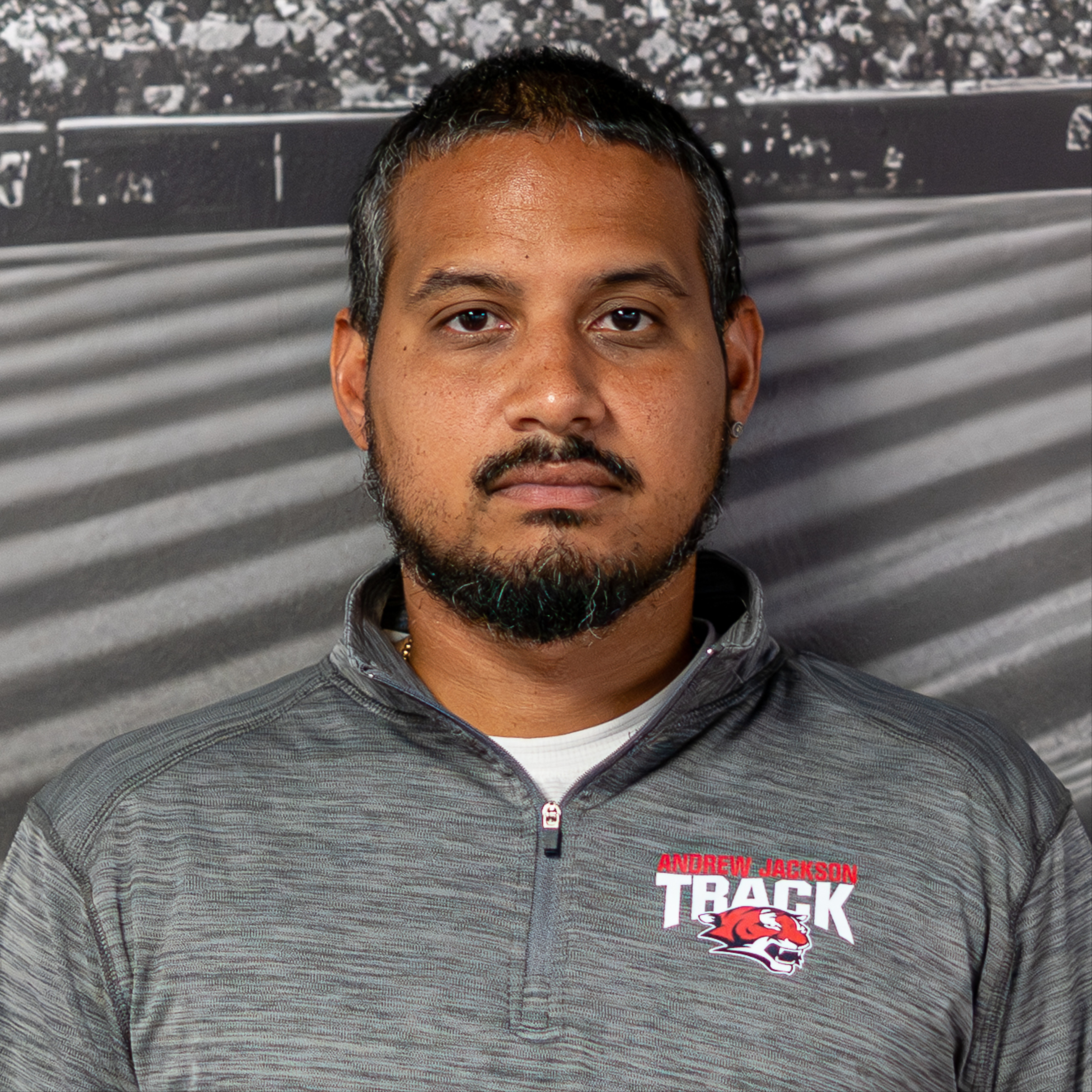 A man stands in front of a large photo of a sports field. He wears a gray jacket with the word "TRACK" printed on it.
