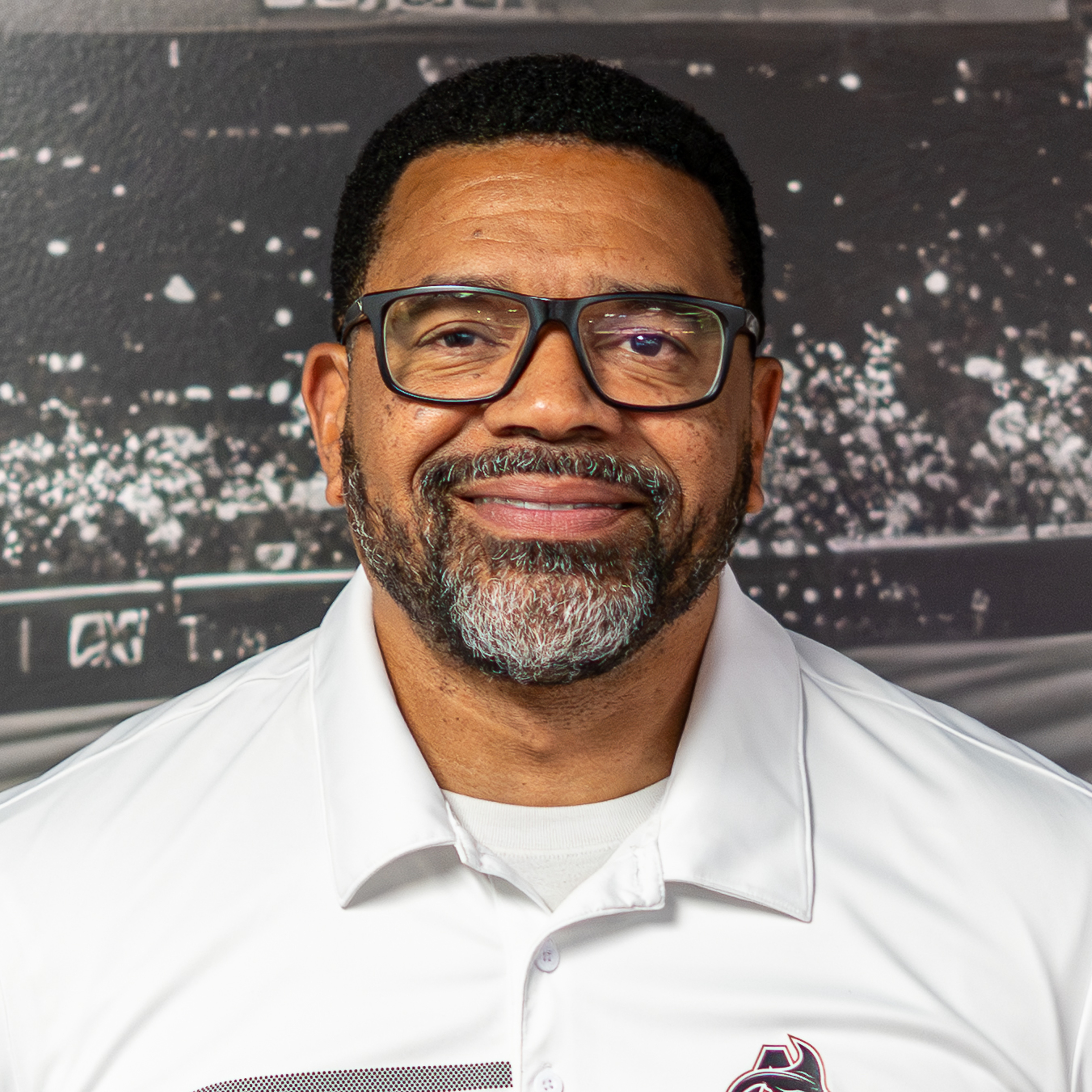 A man wearing glasses and a white polo with a logo smiles against a blurred sports stadium background.