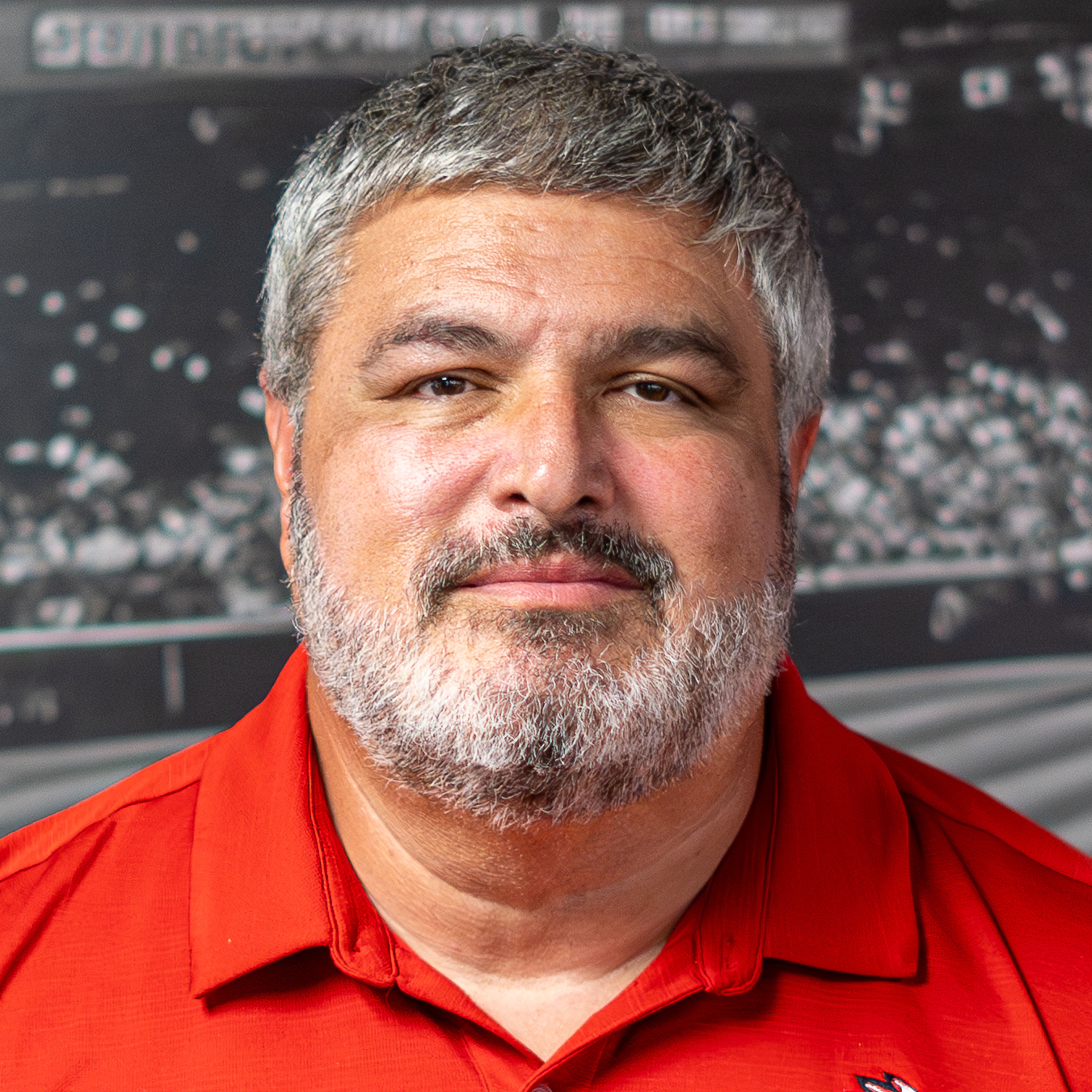 A man with gray hair and a beard wearing a red shirt. Background shows a stadium.