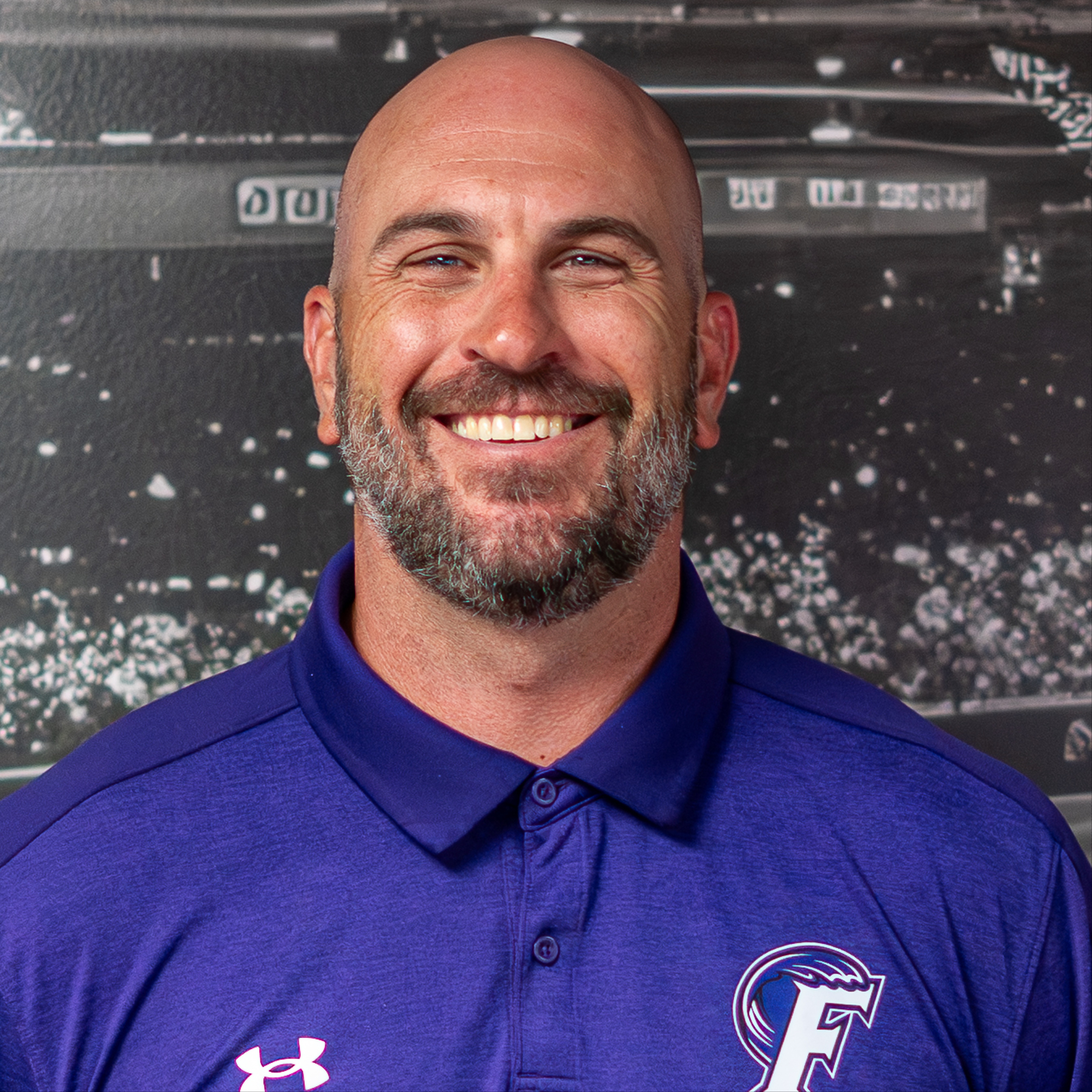 Man with bald head, beard, and blue polo shirt smiles in front of a sports arena.
