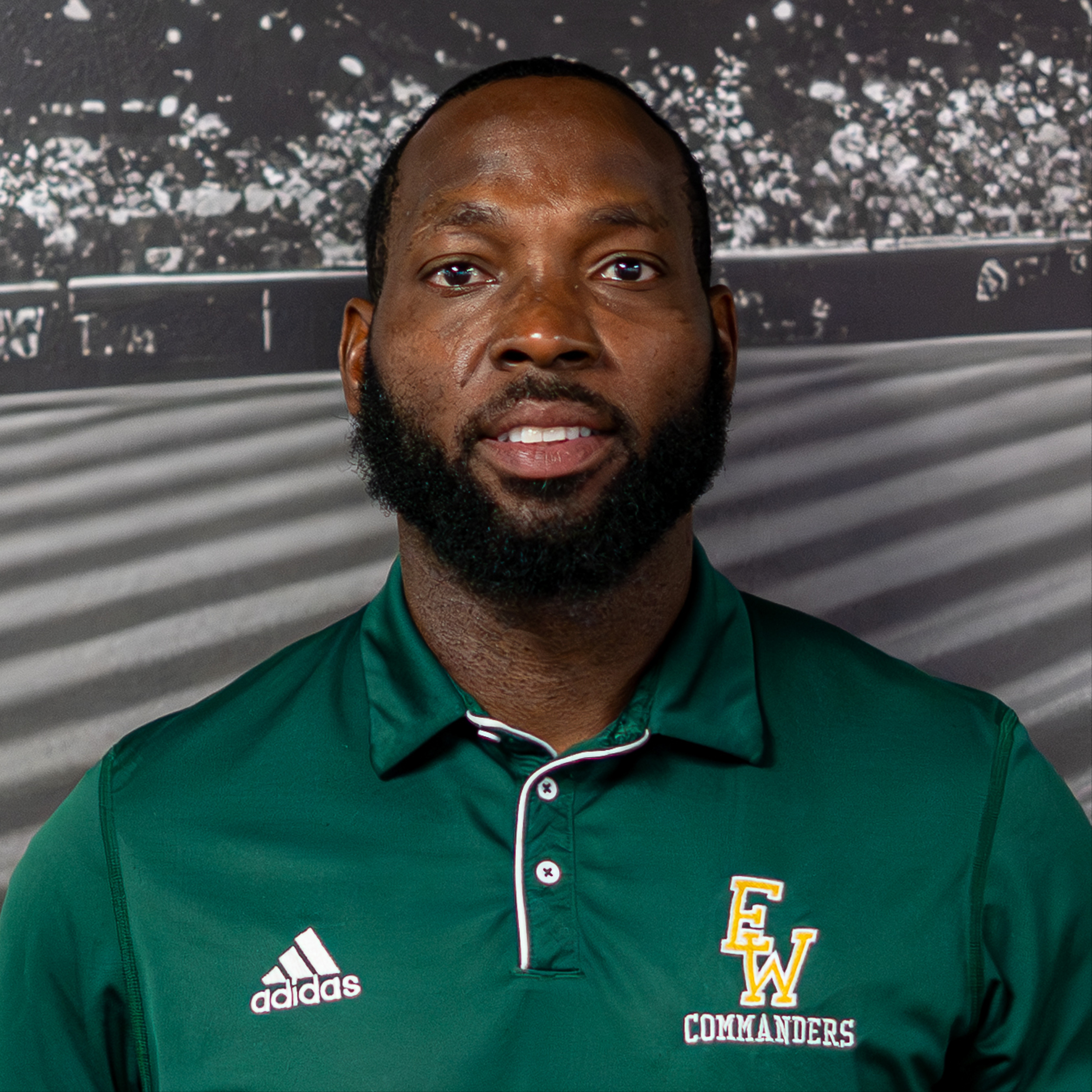 Man with a beard, wearing a green Adidas polo shirt with "EW" and "COMMANDERS" on it, against a blurred sports stadium background.