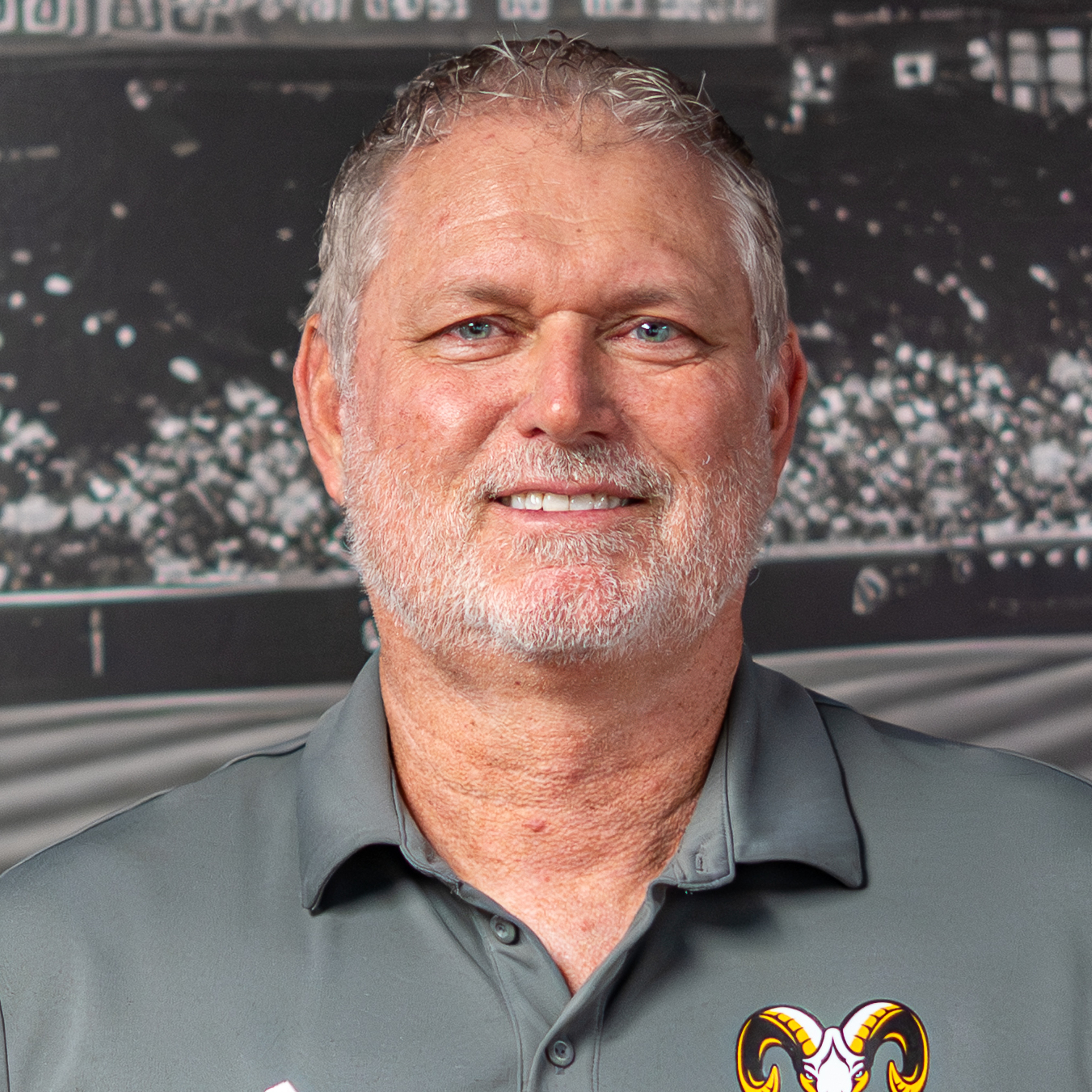 Man in gray polo with logo, white hair, beard, and mustache standing against sports arena backdrop.
