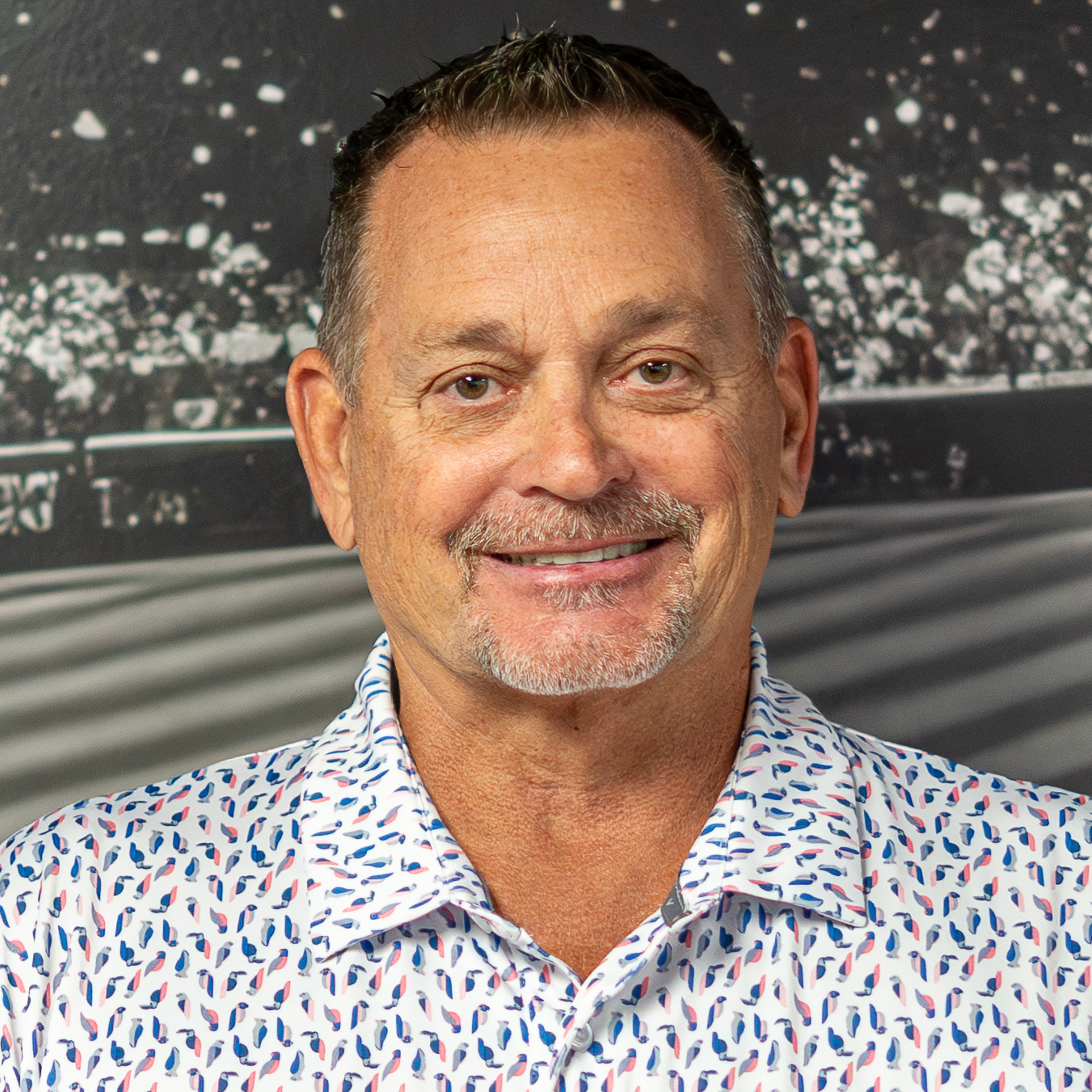Man smiling, wearing a white shirt with a blue and red pattern, against a blurred background with a racing track.