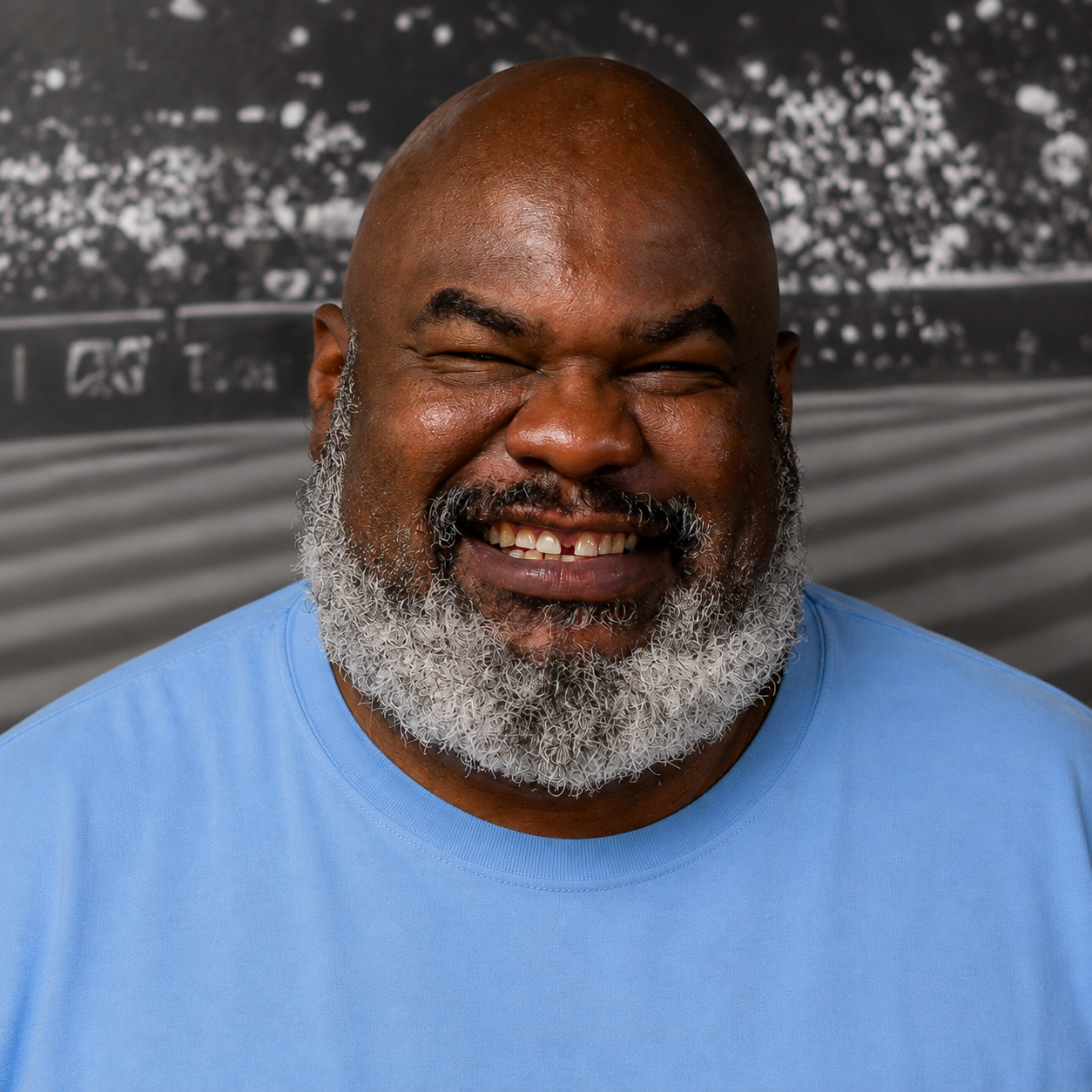 A man with a beard and smile wears a blue shirt against a blurred background with a track.