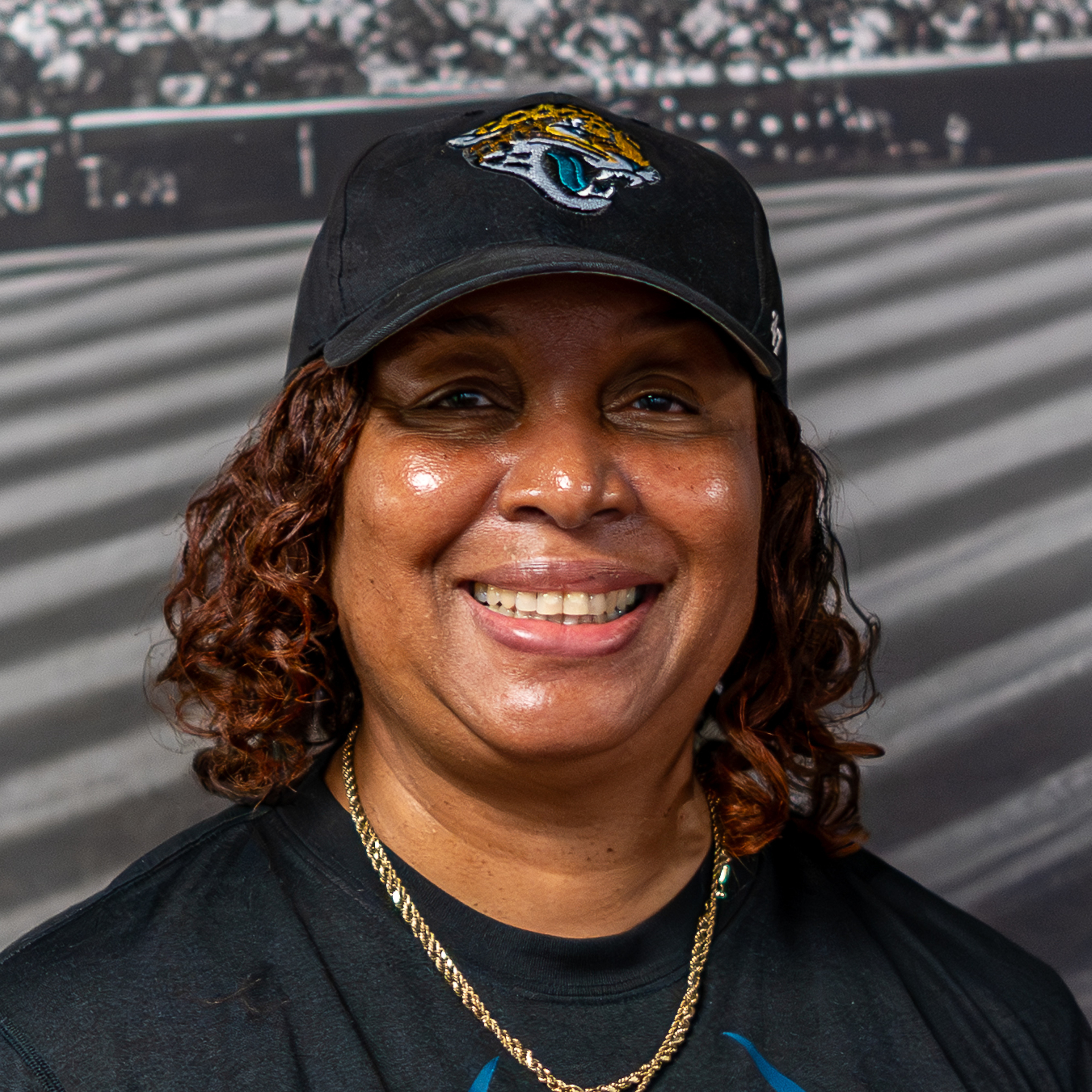 Woman wearing a black hat with a team logo, smiling at the camera. Background shows a blurred stadium scene.