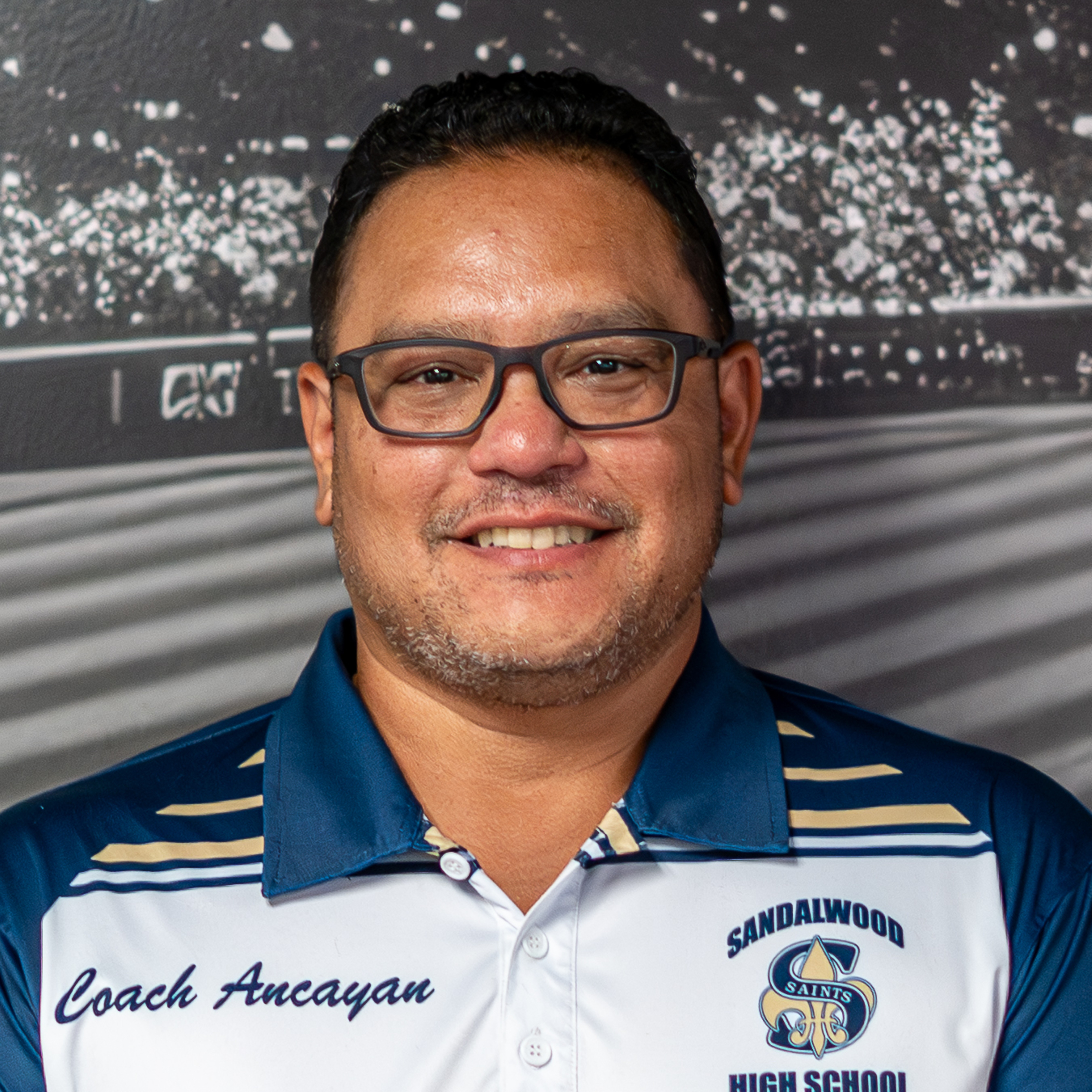 A smiling man with glasses wears a blue and white polo shirt that says "Coach Ancayan" and "SANDLAWOOD HIGH SCHOOL".