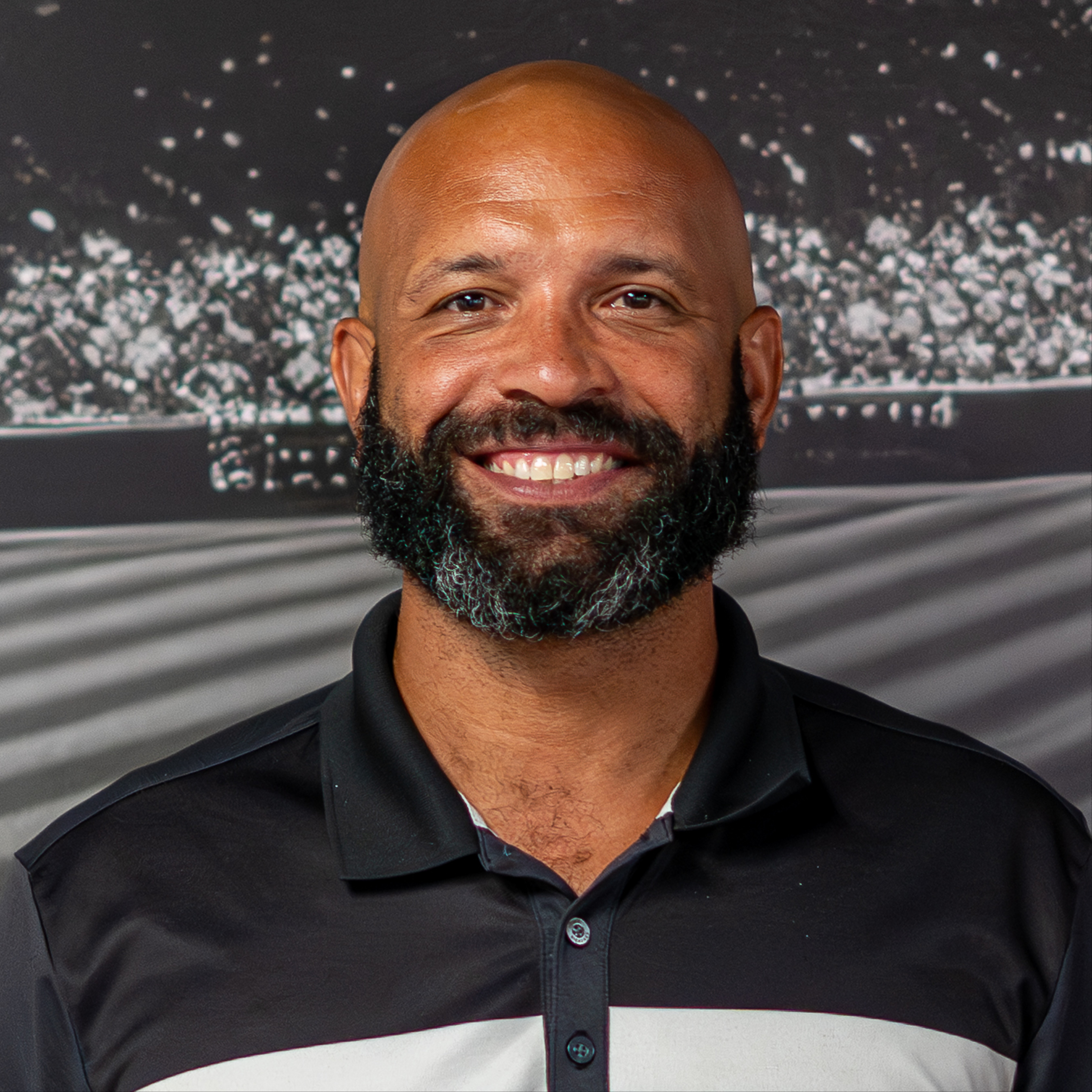 A man with a beard stands smiling. He wears a black and white polo. A sports field is behind him.