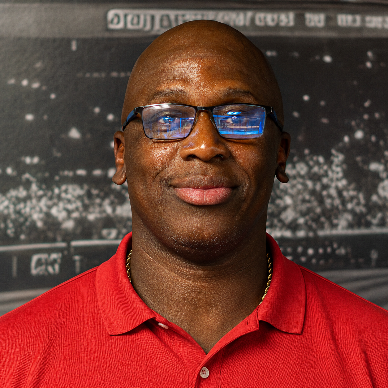 Man in a red shirt, wearing glasses, standing in front of a blurred stadium background.