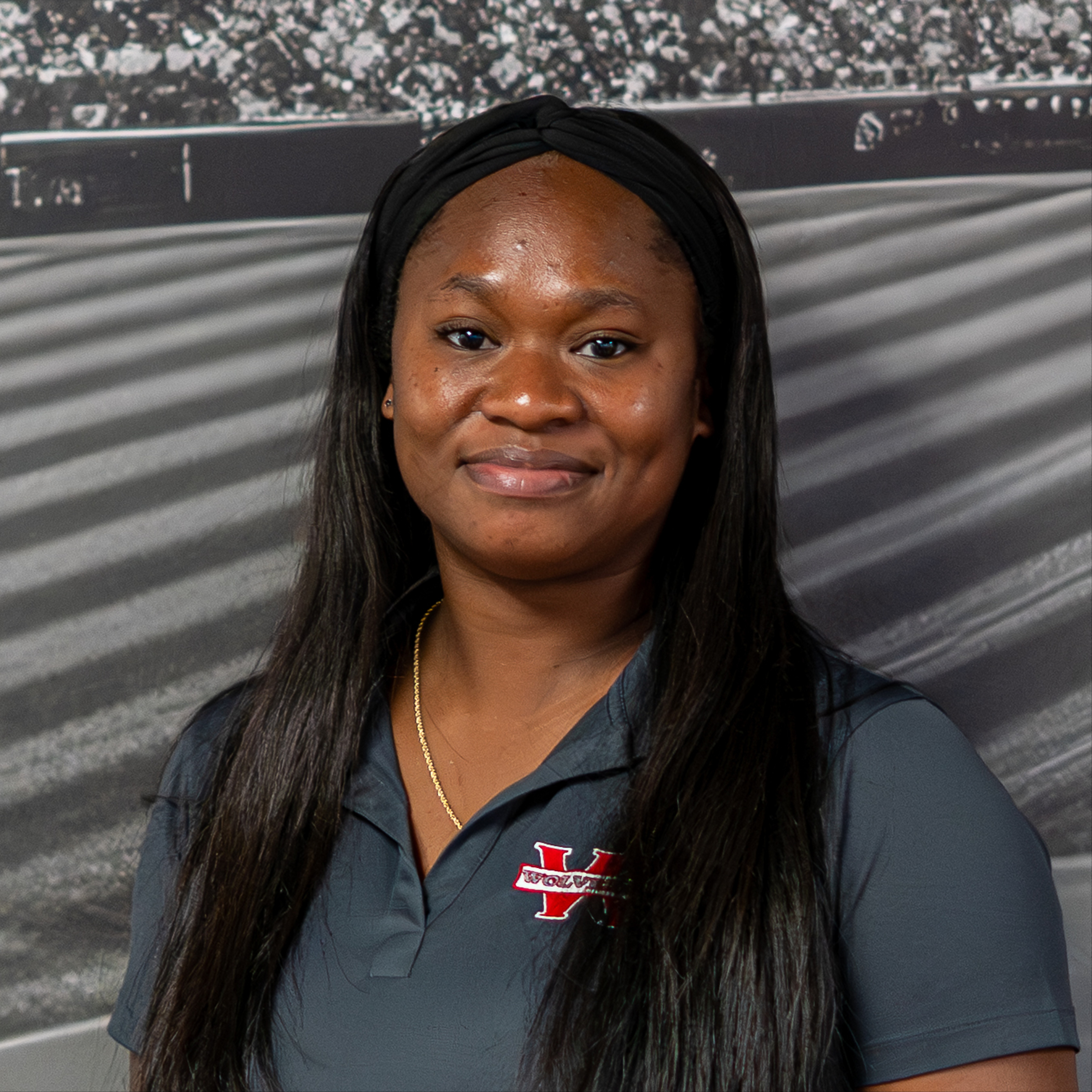 Woman with long hair wearing a gray polo with red logo smiling in front of a blurred stadium background.