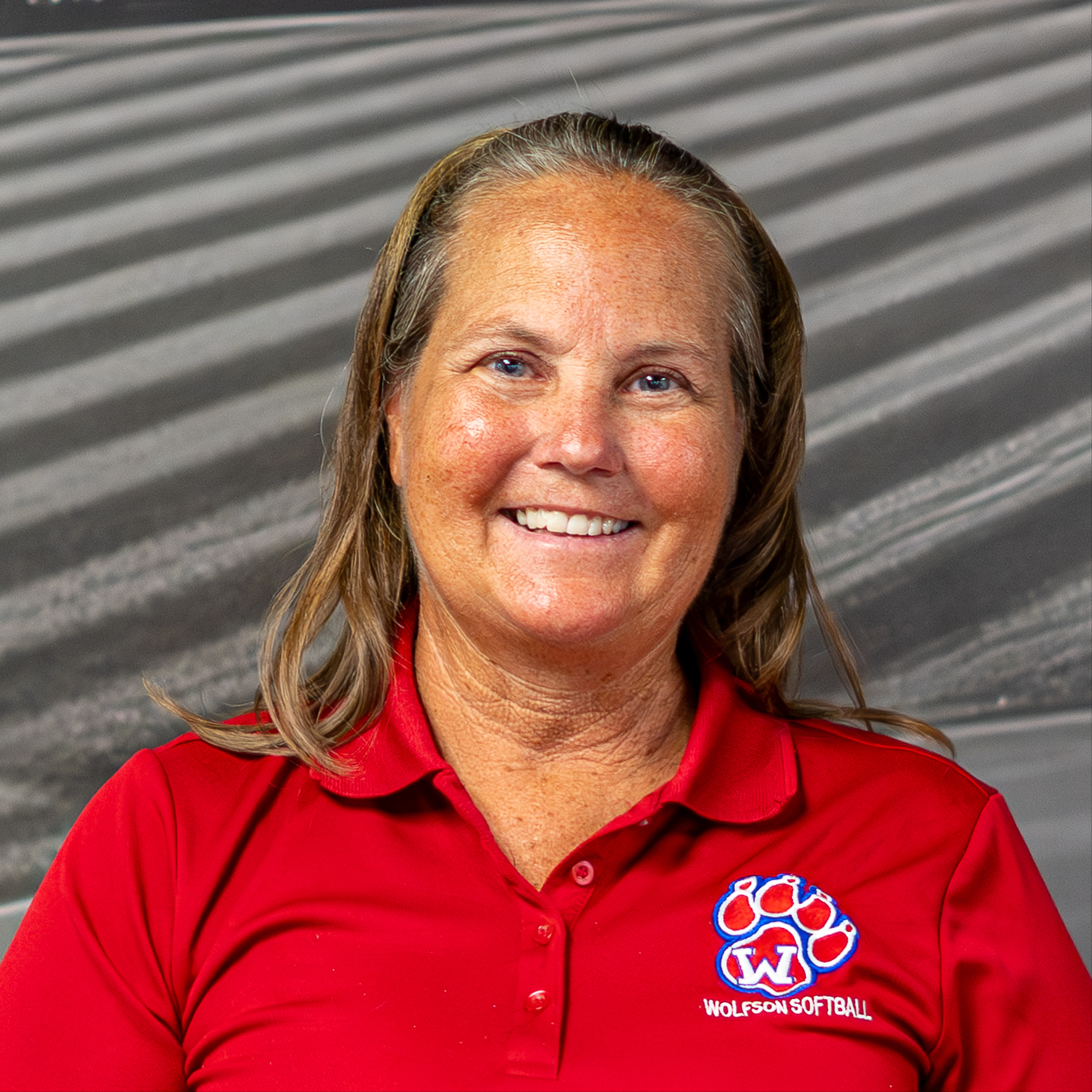 A woman with blonde hair wearing a red shirt with a logo is smiling in front of a gray background.