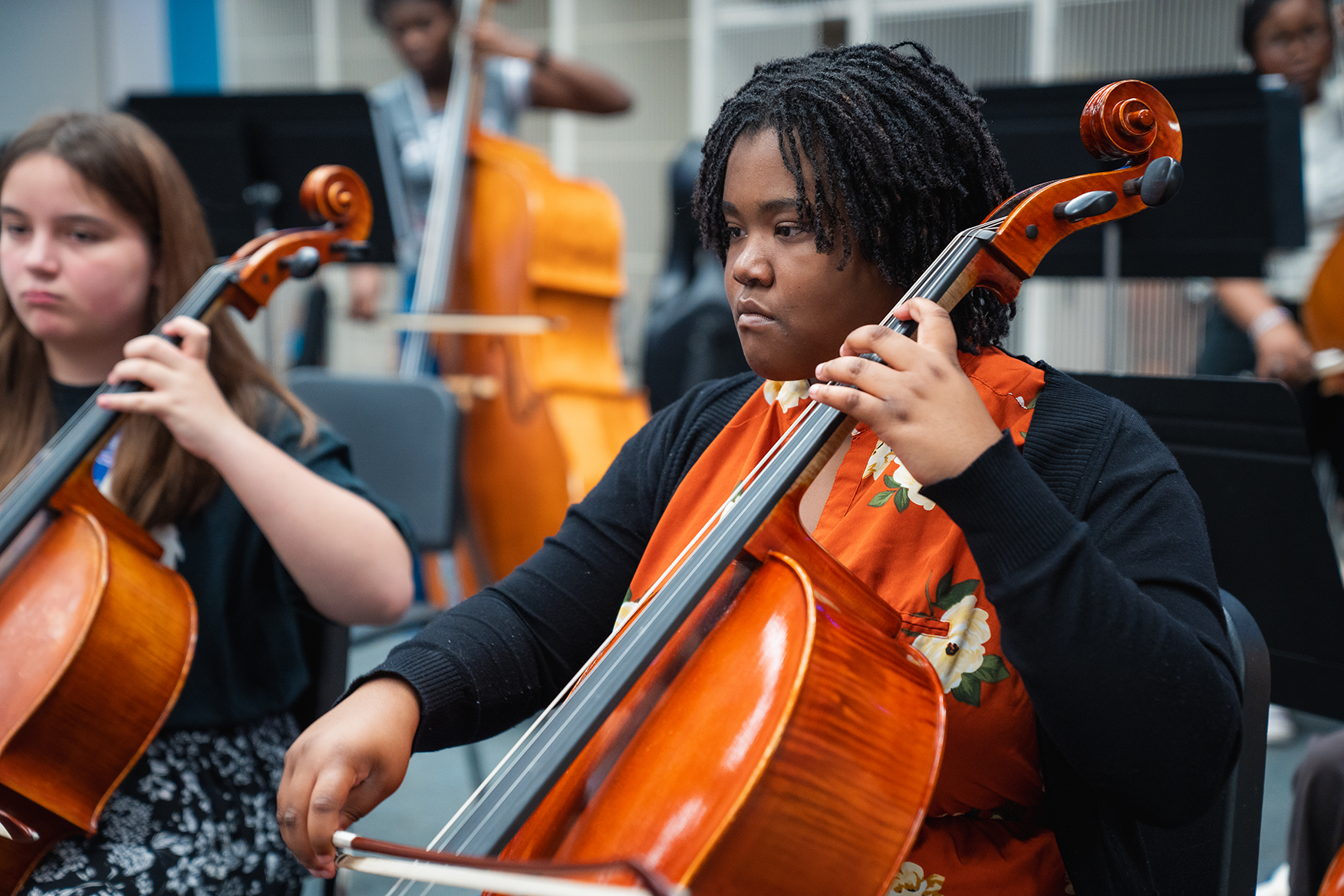 Two people play cellos in a classroom. One is seated and focused on the instrument. The other stands beside them.