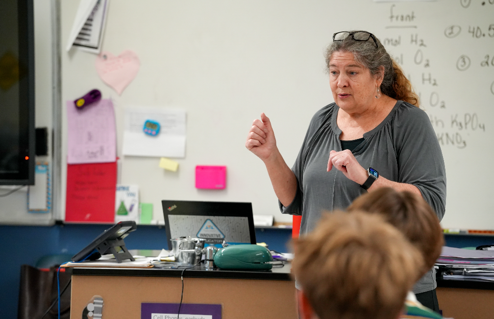A woman teaching, standing at a desk, gesturing with her hands in a classroom with students.