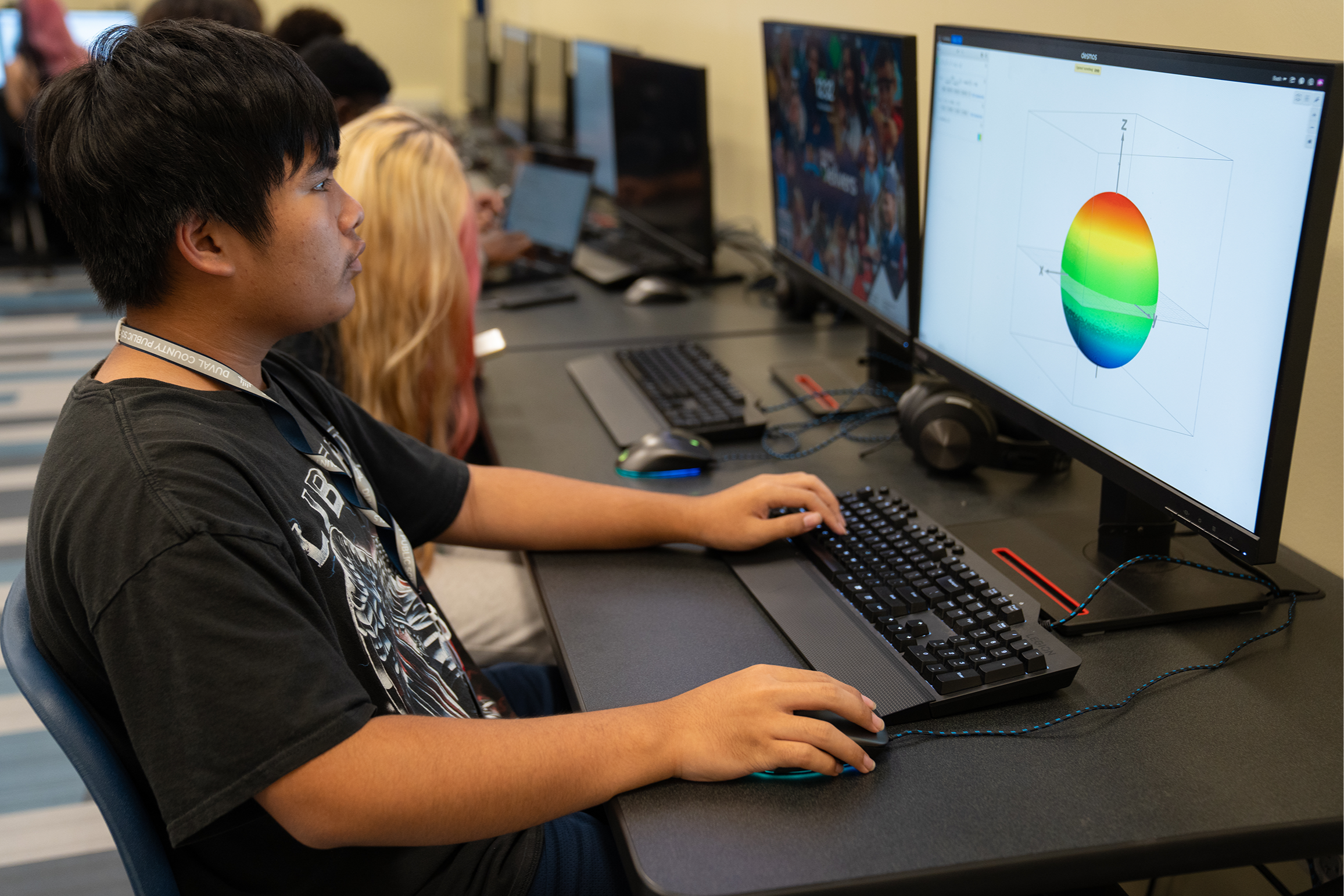 A student using a computer, sitting at a desk with two screens, a keyboard, and a mouse.