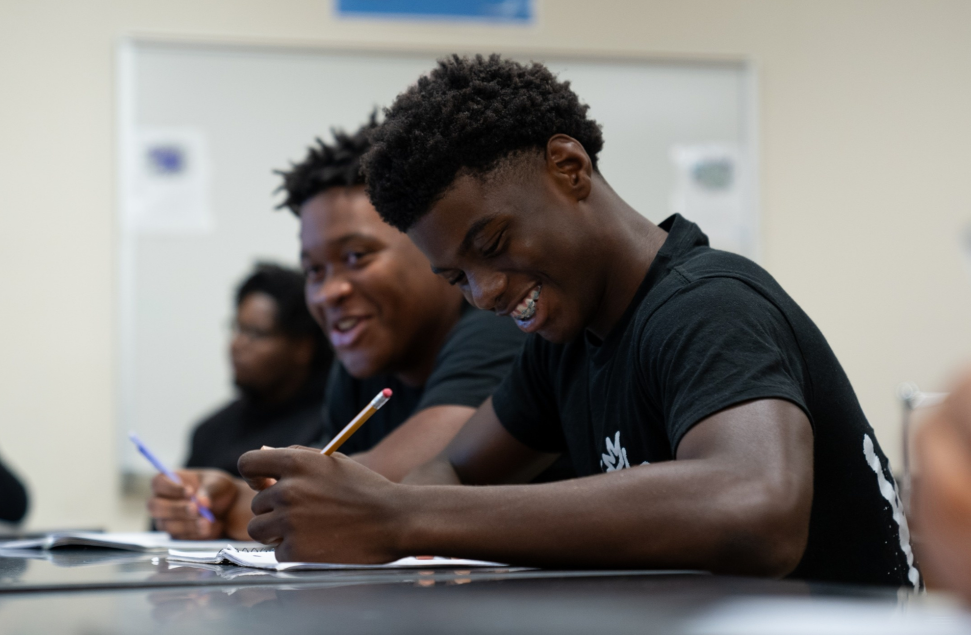 Two students sit at a table, smiling and working. One holds a pencil, the other a pen.