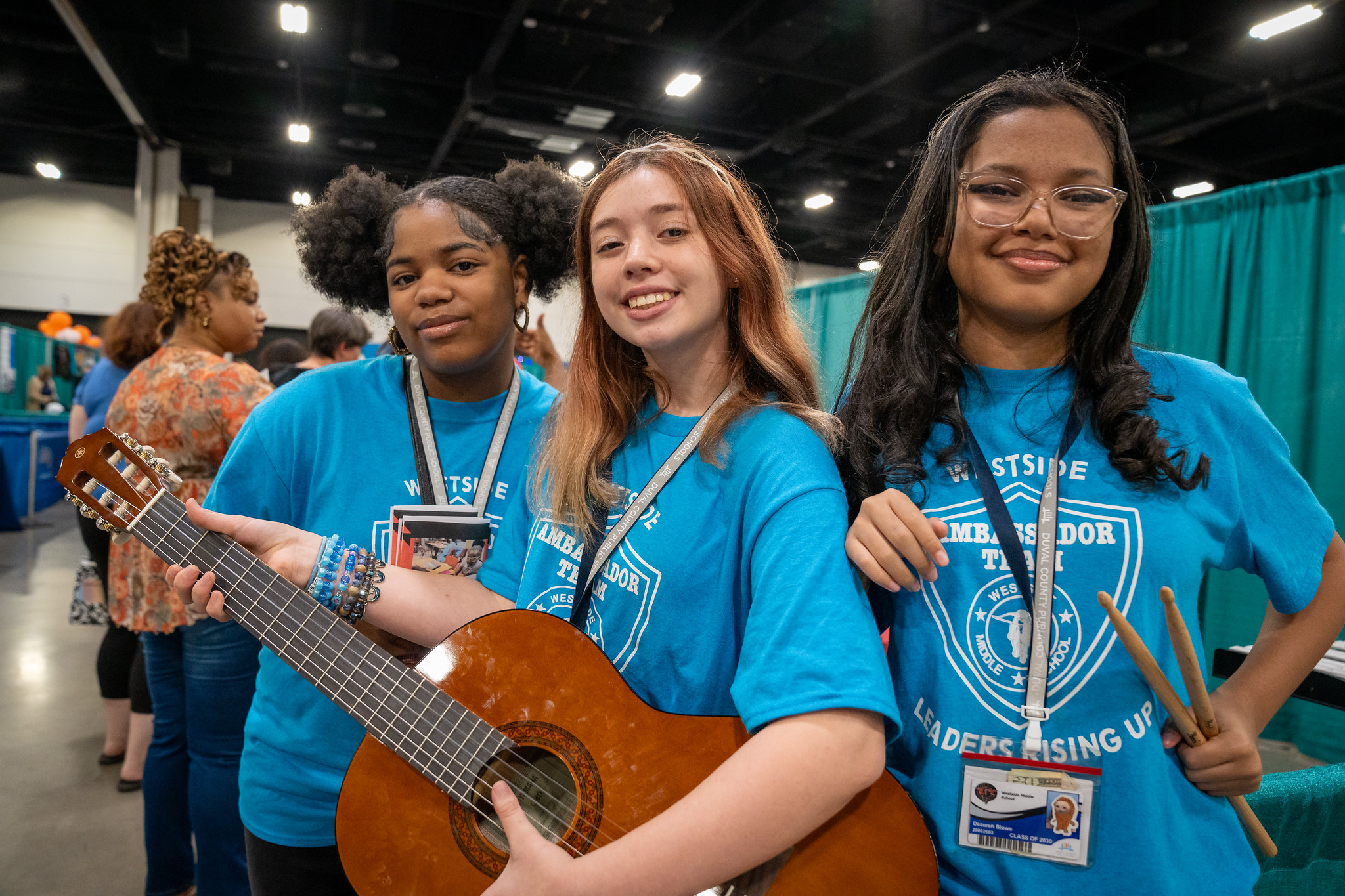 Three women in blue shirts hold a guitar and drum sticks. One woman smiles, and the other two pose.