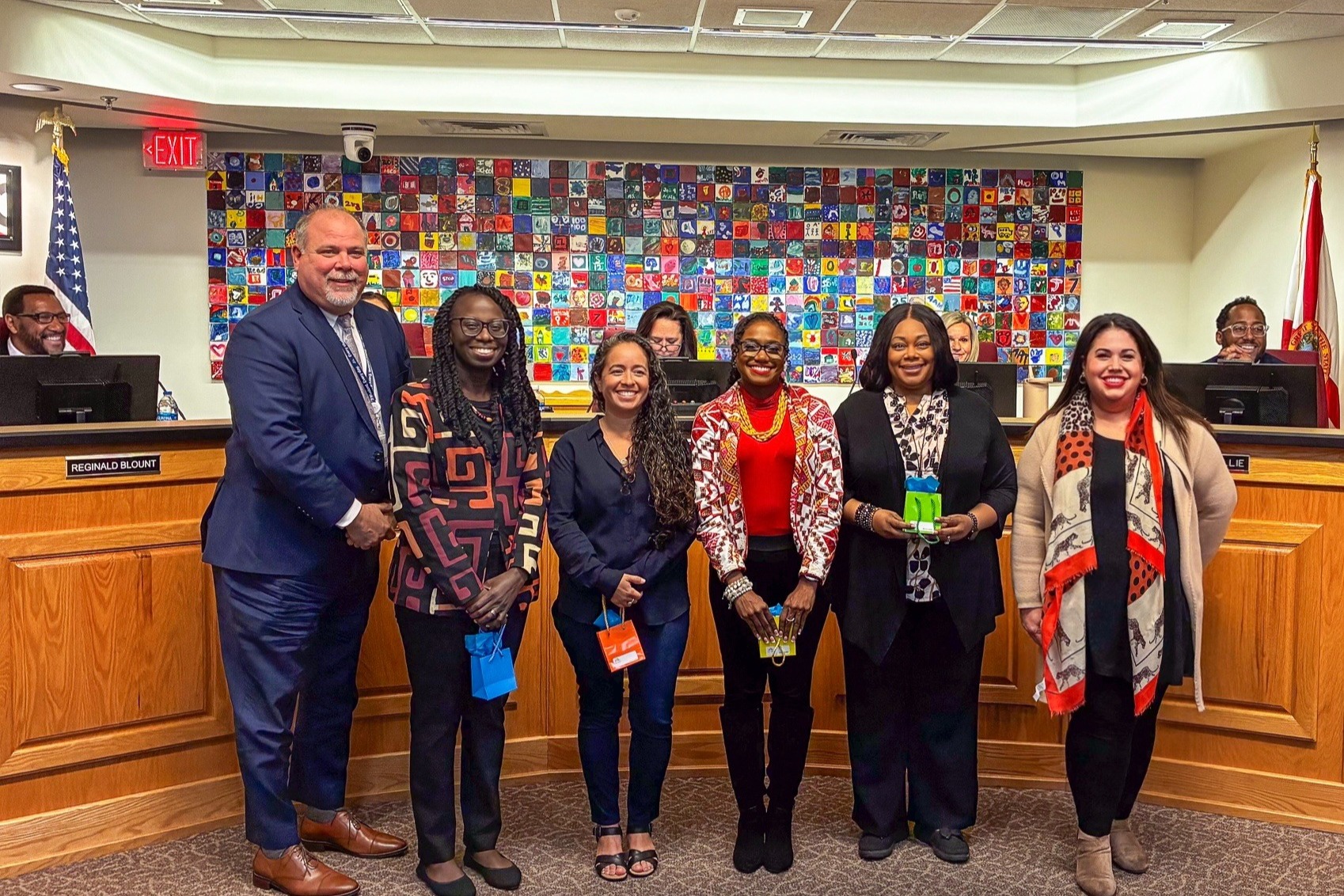 A group of people stand in front of a desk, including a man in a suit. Some wear coats and scarves. A wall behind them is colorful.