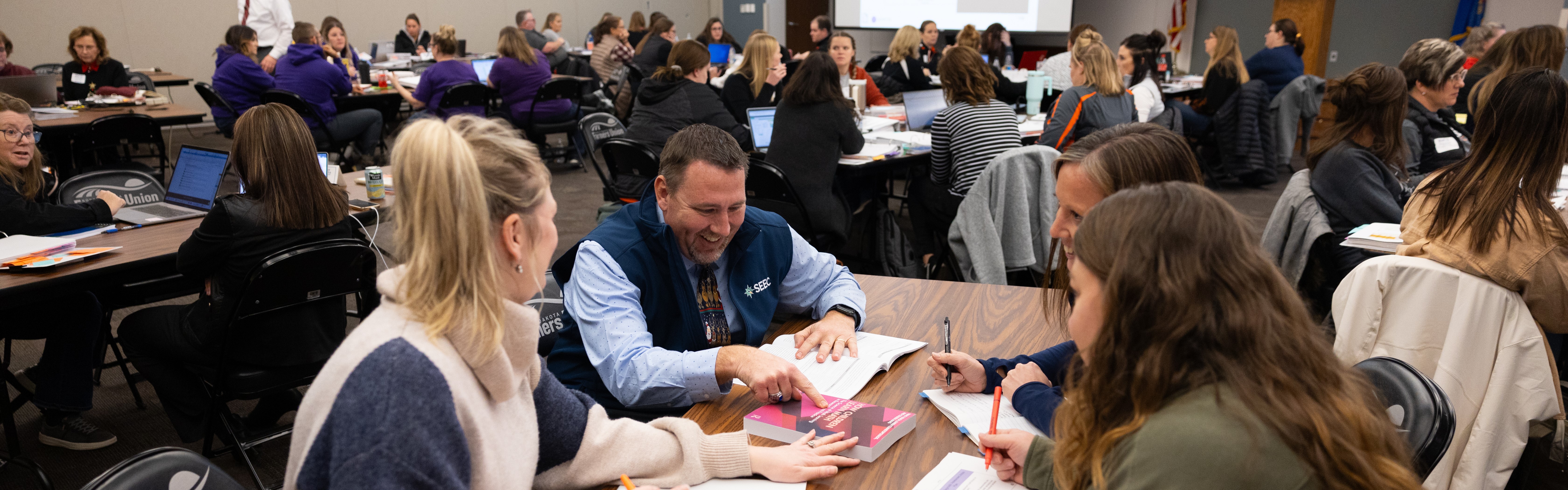 educators sitting at tables working on sheets of paper pointing at a book at the NDMTSS Training