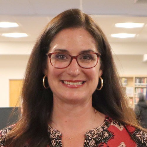 A woman in glasses standing in front of a library, looking at the camera with a smile.