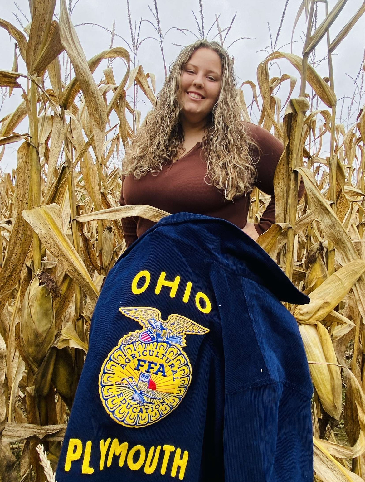 A woman standing in a corn field wearing a jacket, surrounded by tall stalks of corn.