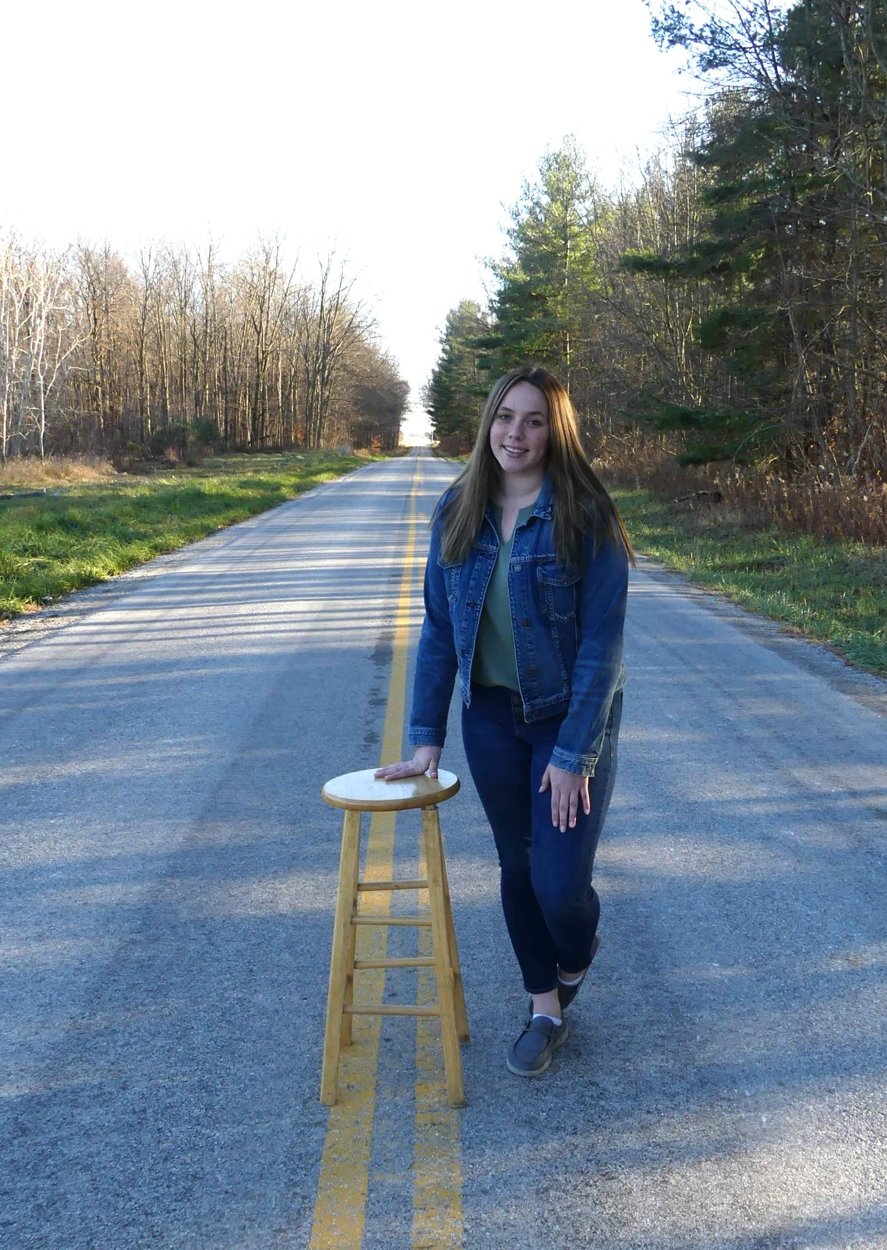 A woman standing next to a wooden stool on a road, under an open sky.
