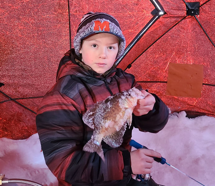 A boy joyfully holds a fish inside a cozy snow-covered tent, surrounded by a serene winter landscape.