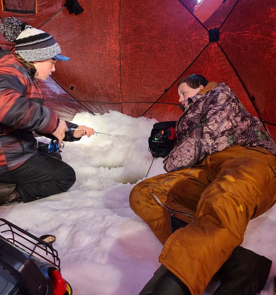 Two men sitting in a snow-covered tent, surrounded by a white winter landscape.